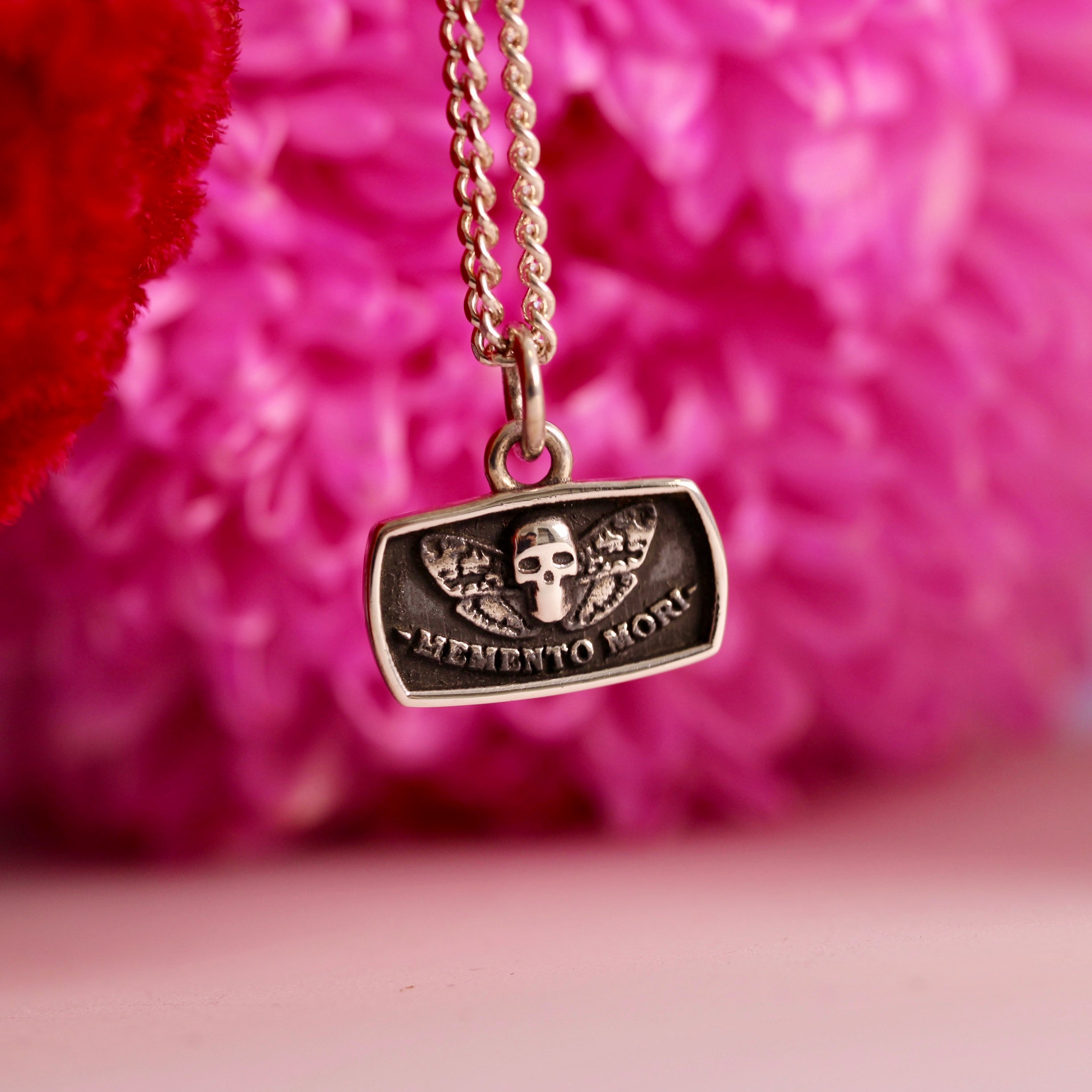 A silver death moth memento mori necklace with a rectangular pendant featuring a skull and crossbones design, mounted on a chain with a pink floral background.