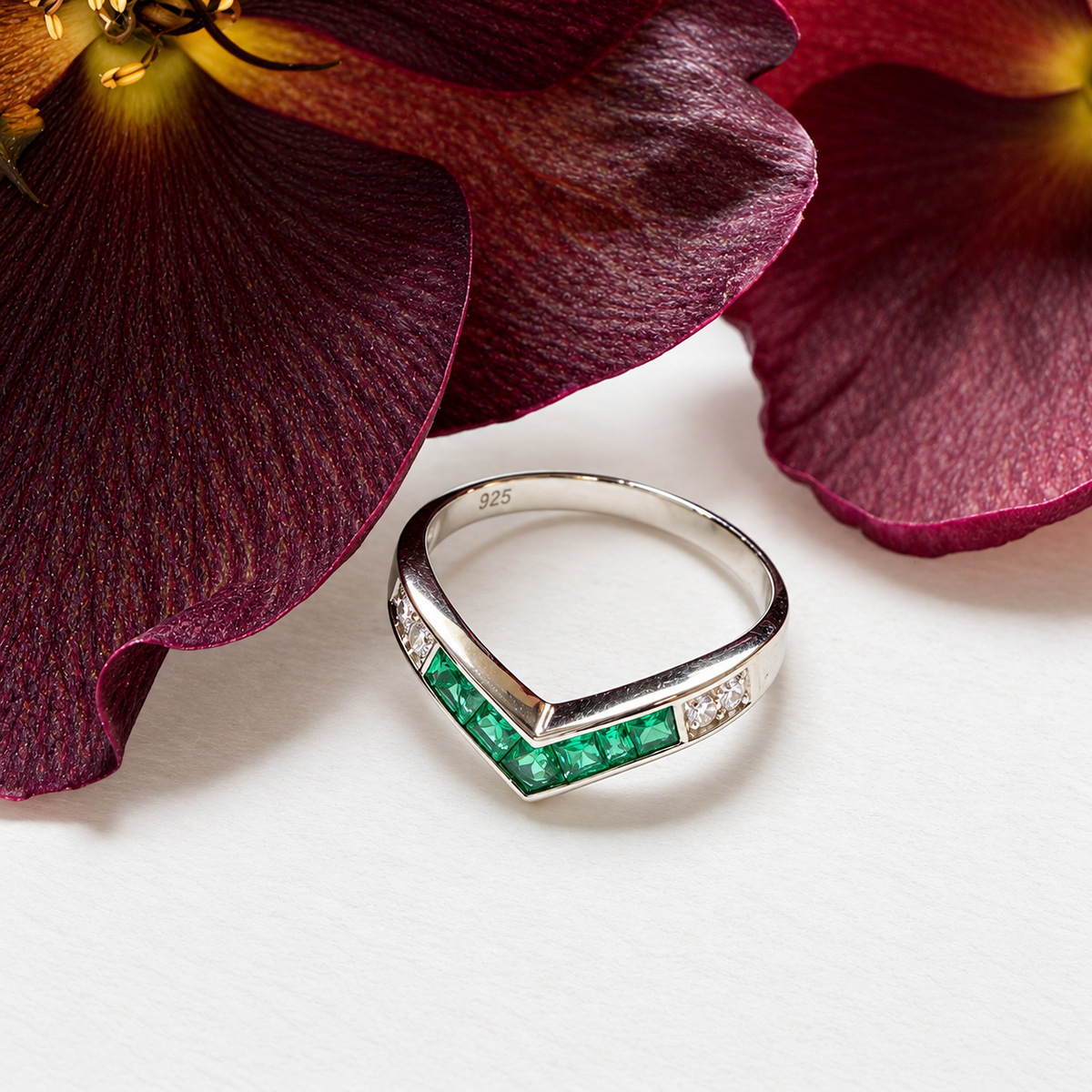 A silver ring with a chevron design featuring a row of green emerald-cut stones and small clear stones on the sides, styled on a white surface with large dark burgundy flower petals in the background.
