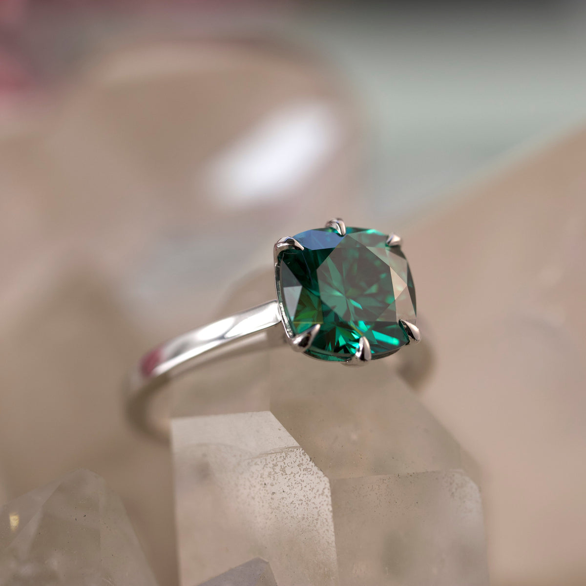A white gold ring with a cushion-cut green gemstone, resting on clear crystal blocks against a soft, blurred background.