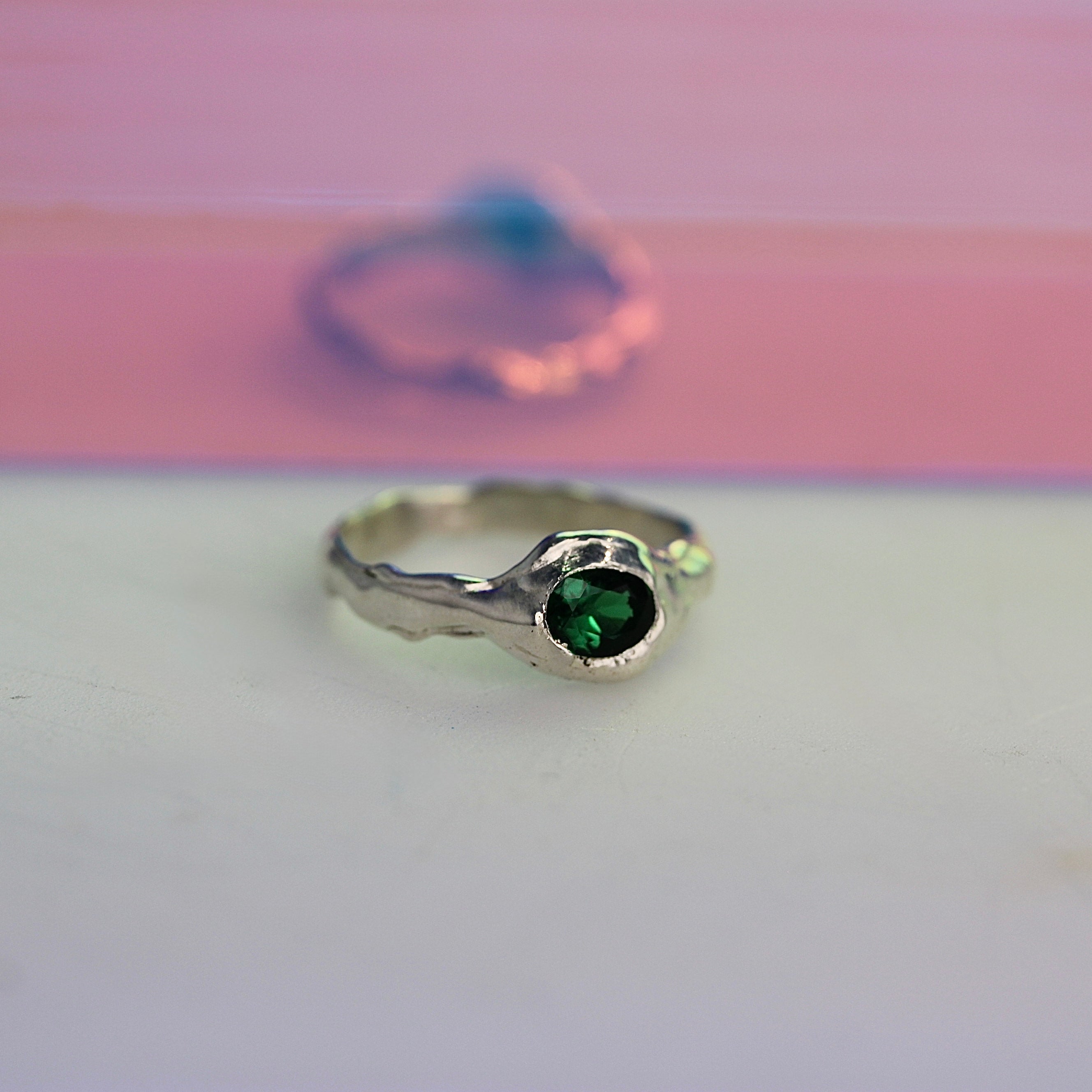 A unique, handcrafted silver ring with a bold textured design and an inlaid green oval nano gemstone, displayed against a pink and white background.