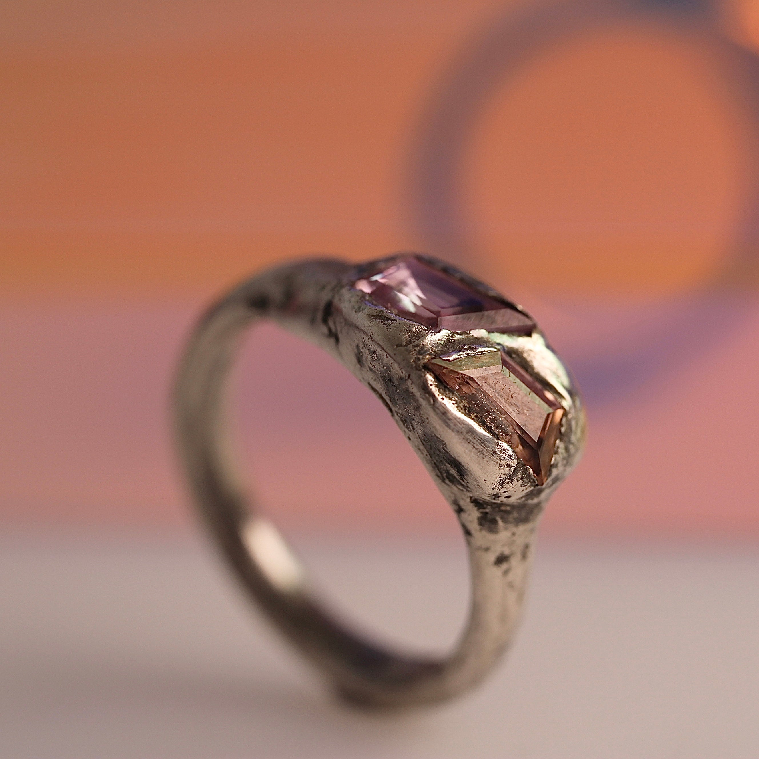 A handcrafted silver ring with two pink sapphires set in a textured design, photographed against an orange and pink background.