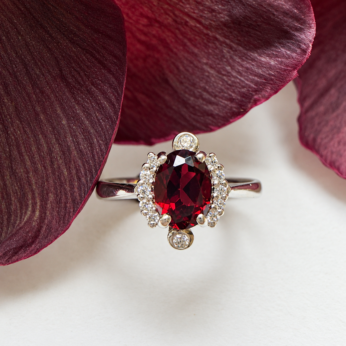 An oval-cut red gemstone ring with a halo of small clear stones, photographed close up against white with dark burgundy flower petals.