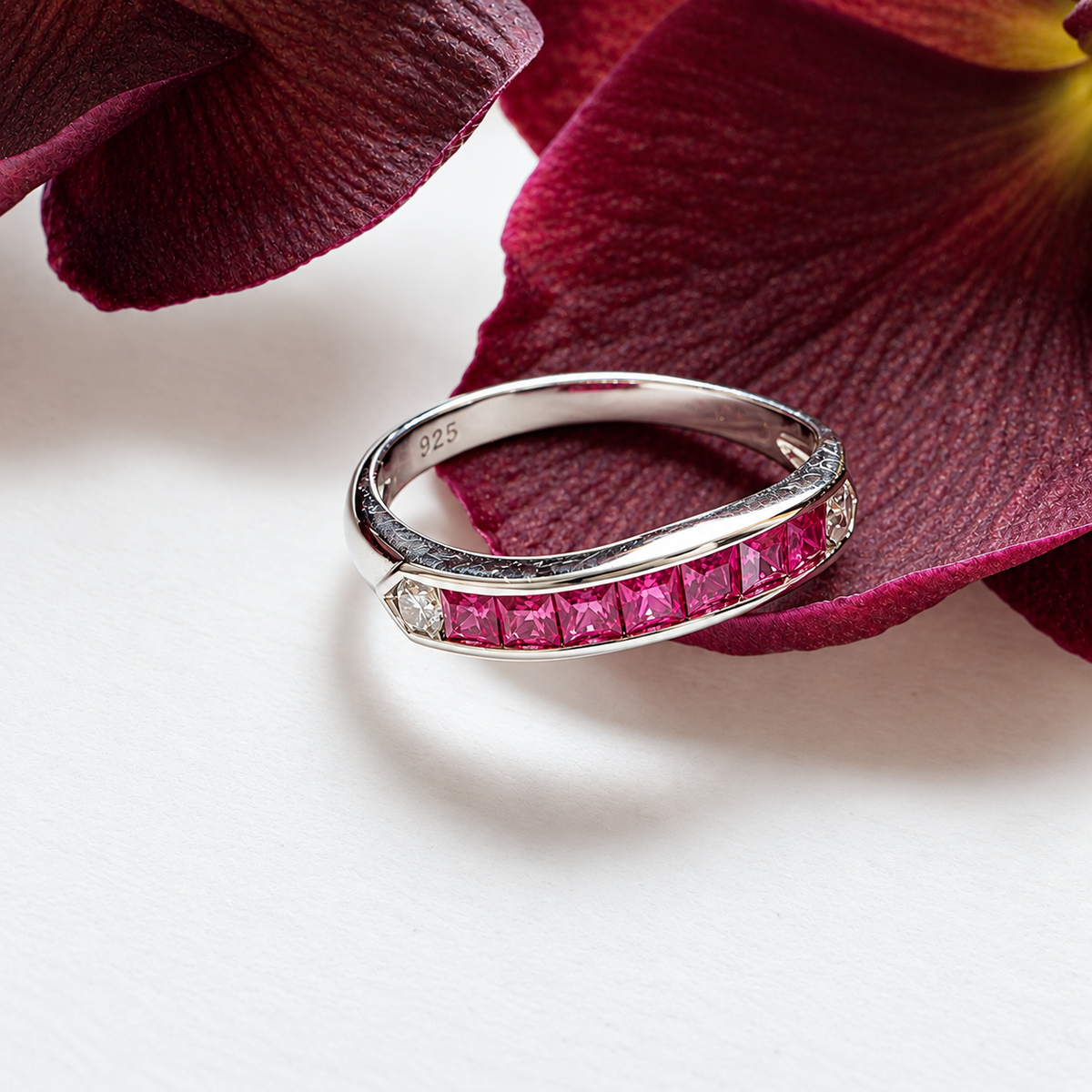 A silver band ring featuring a row of pink gemstones and a small clear stone, photographed on a white surface with dark burgundy flower petals in the background.