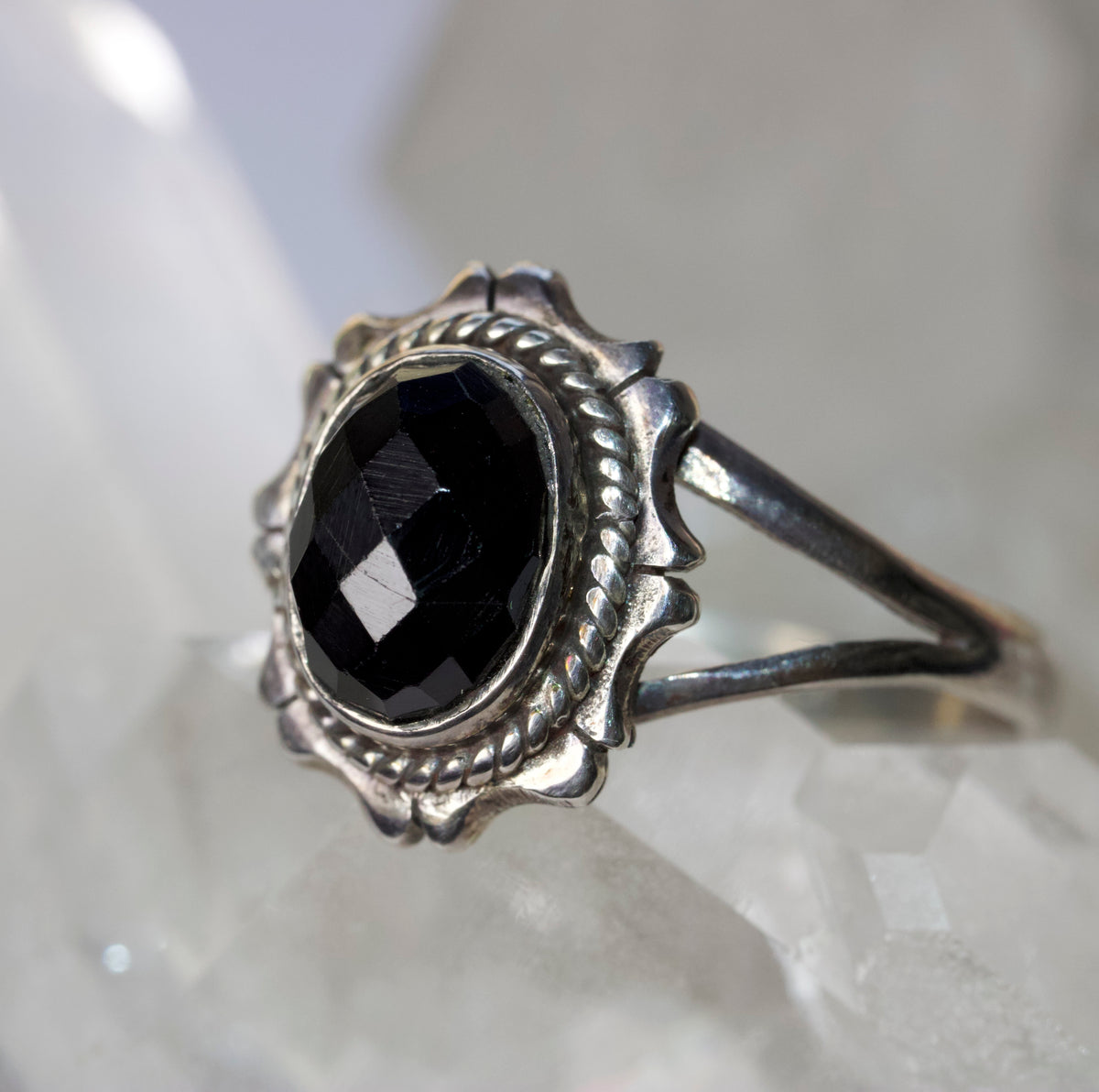 A silver ring featuring a large, faceted black gemstone in a decorative, scalloped circular setting, resting on a white crystal display.