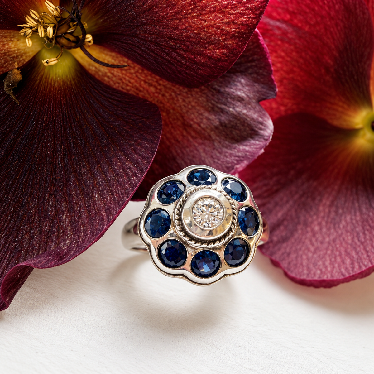 A silver ring with a floral cluster setting featuring round blue sapphires and a central clear diamond, styled on a white surface with large dark red flowers in the background.
