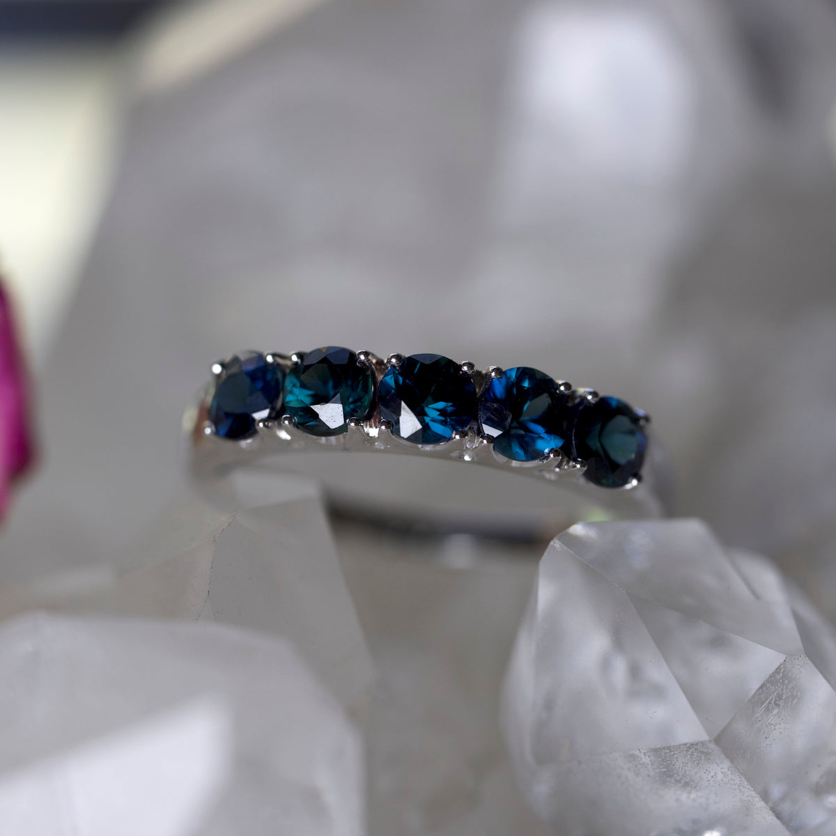 A silver band ring featuring five round-cut partii gemstones, displayed on a bed of white crystal points.