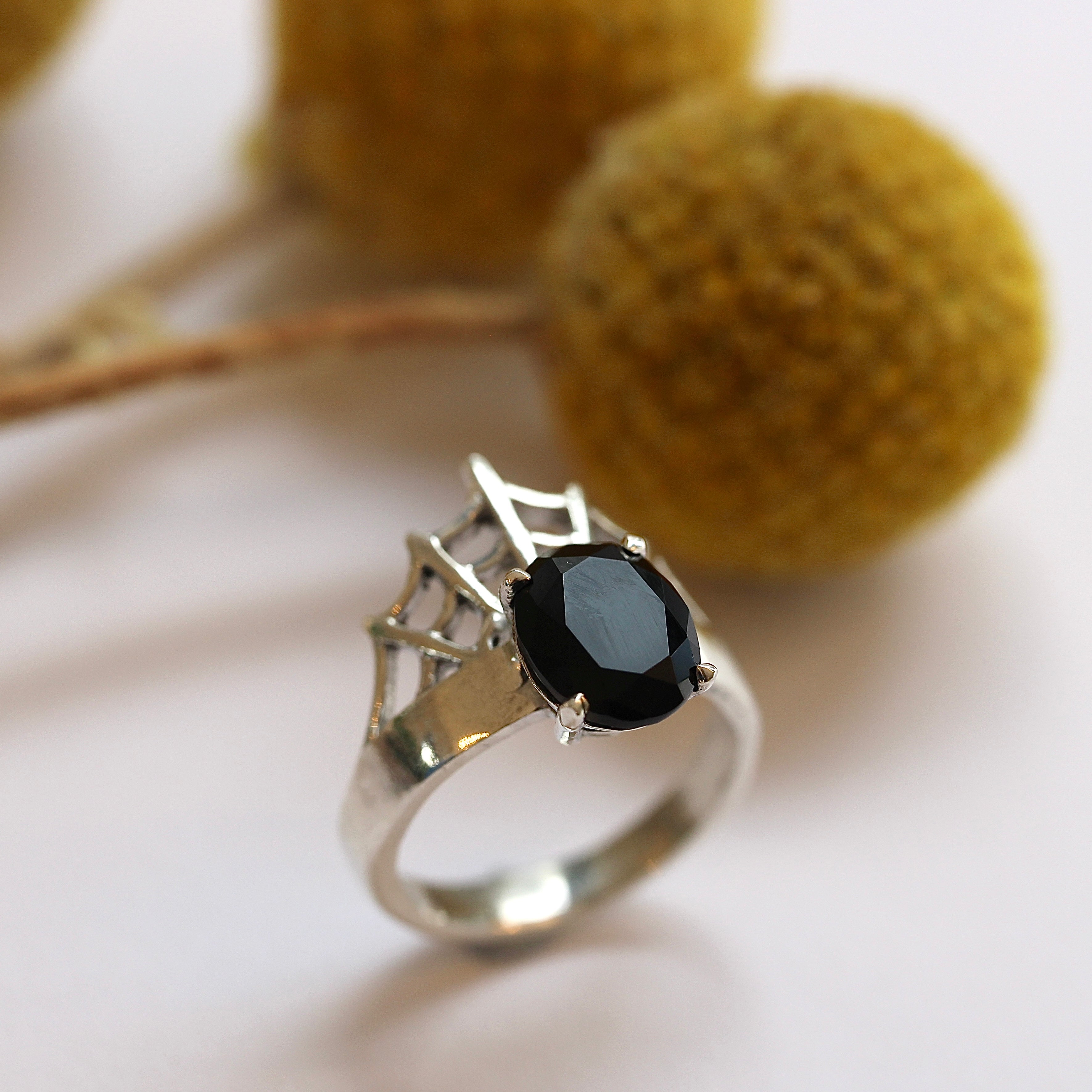 A silver ring with a black oval center stone, featuring a web design on the band, displayed against a neutral background with yellow floral accents.