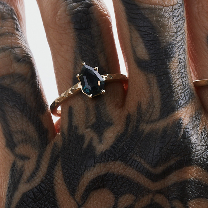 A gold band solitaire ring with a black pearl on a person's finger with tattooed patterns in the background.