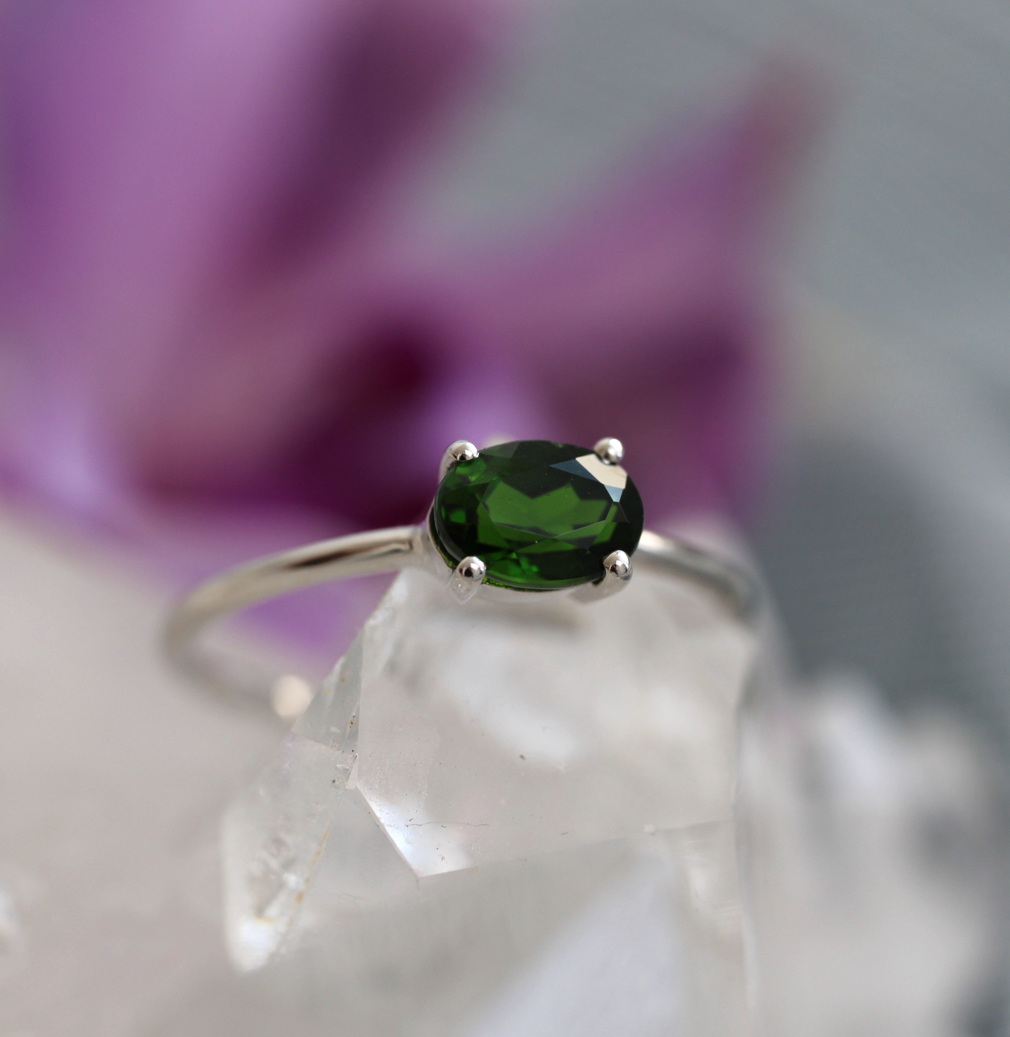 Ring with a green chrome diopsidegemstone on a clear crystal surface with a blurred purple flower in the background