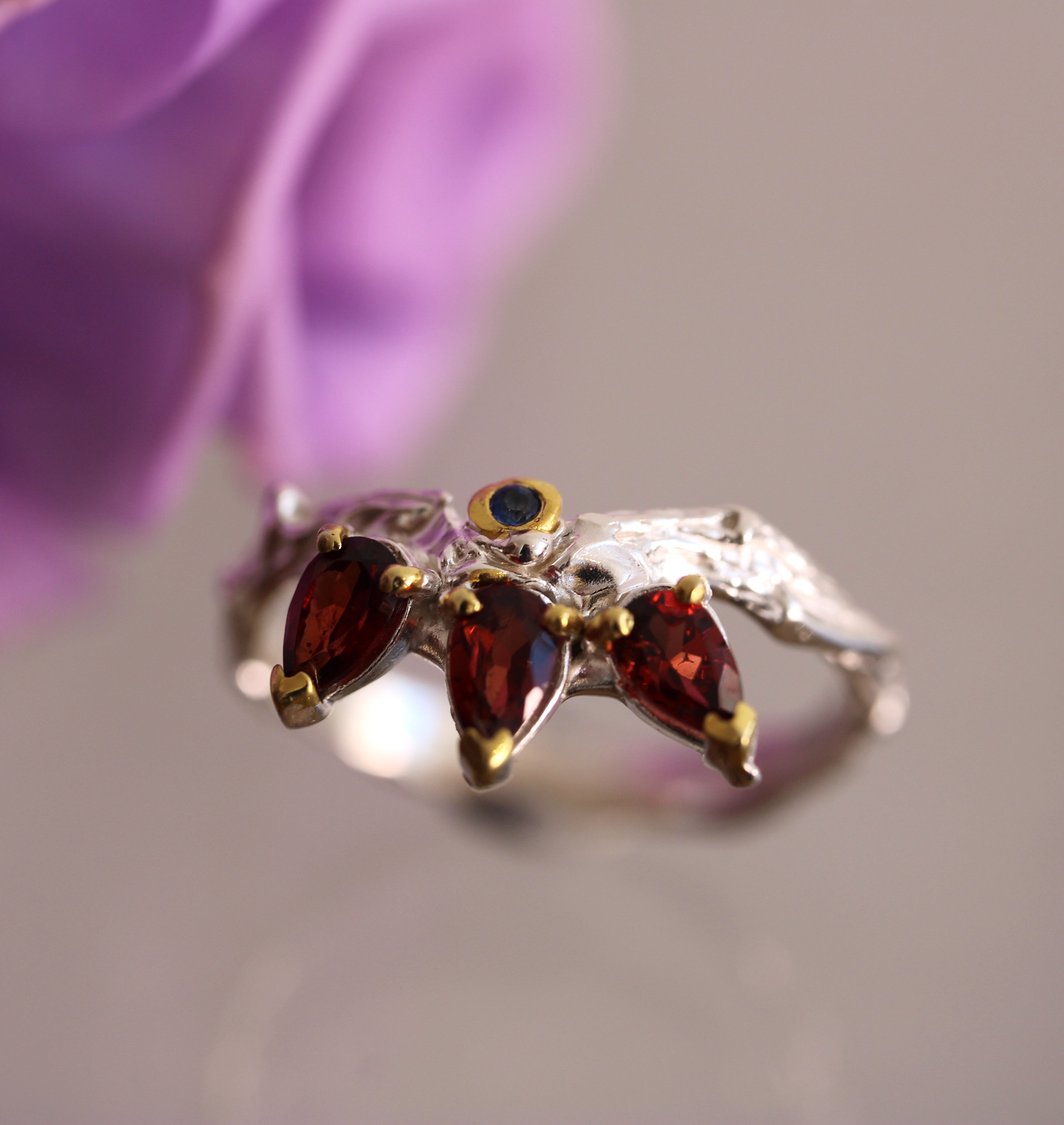 Silver ring with red gemstones against a blurred purple flower background