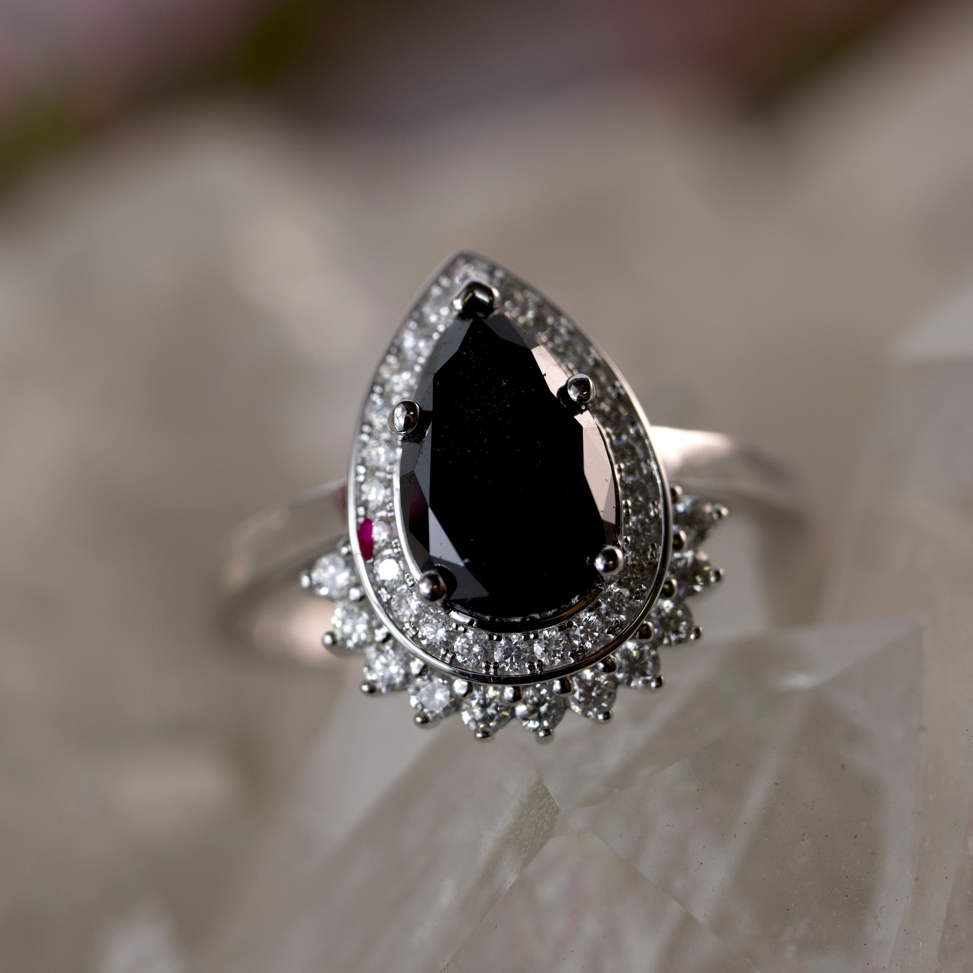 A silver-toned teardrop ring featuring a large black teardrop black diamond surrounded by a halo of small clear stones, displayed on a soft, blurred light beige surface.