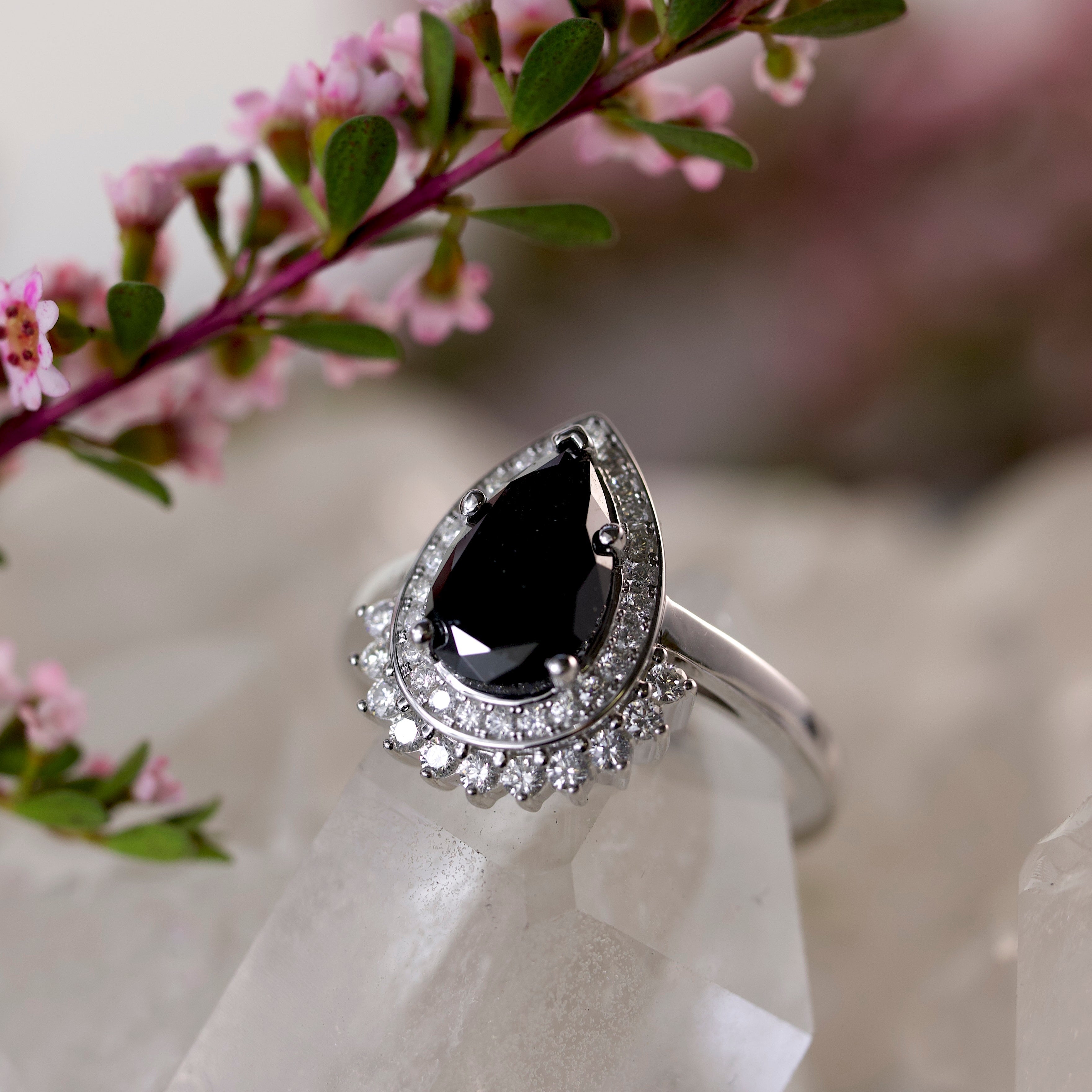 A silver ring featuring a teardrop-shaped black diamond surrounded by a halo of small clear stones, displayed on a clear crystal block with pink flowers in the background.