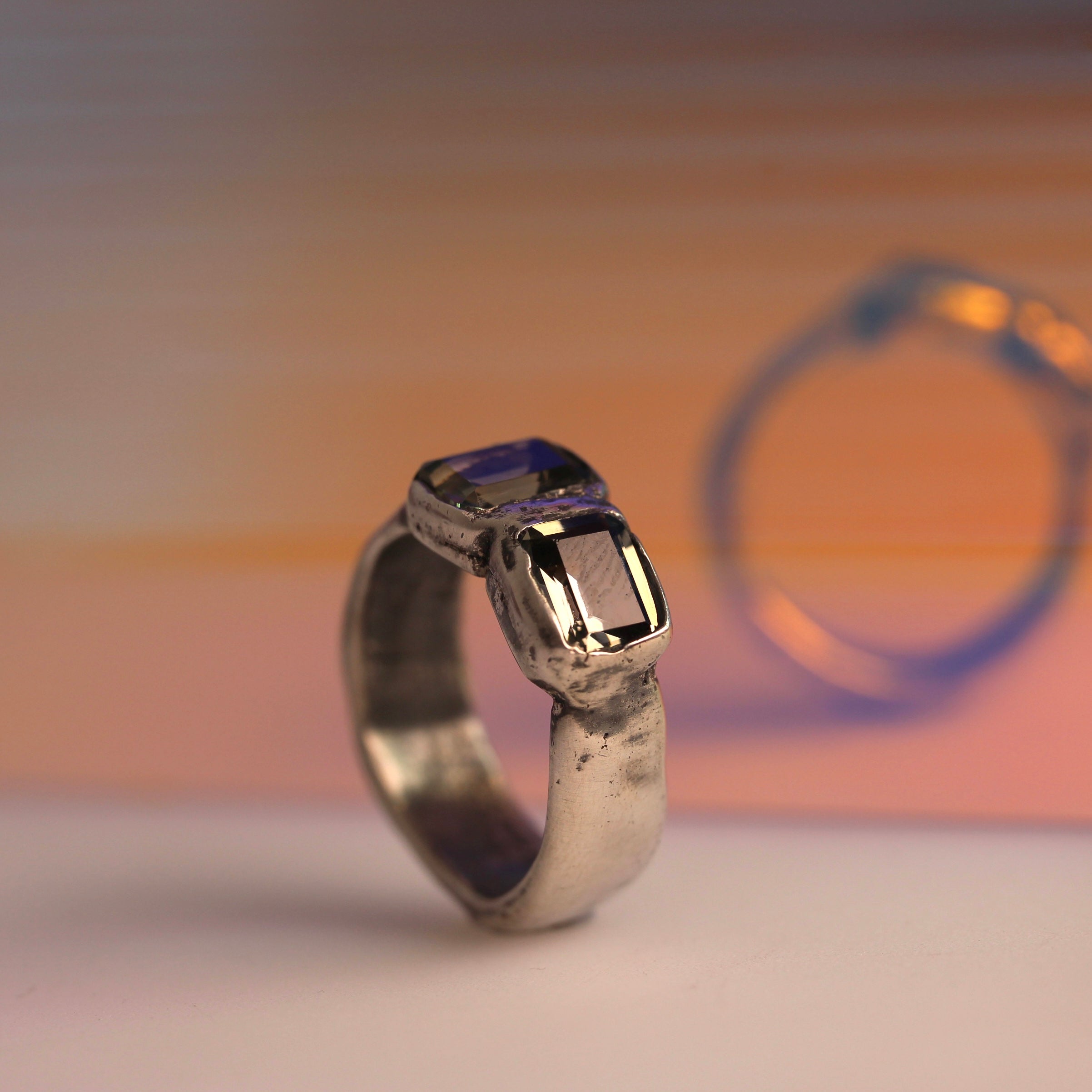 A handcrafted silver ring with two dark grey nano gems set in a textured design, displayed against a warm-toned background.