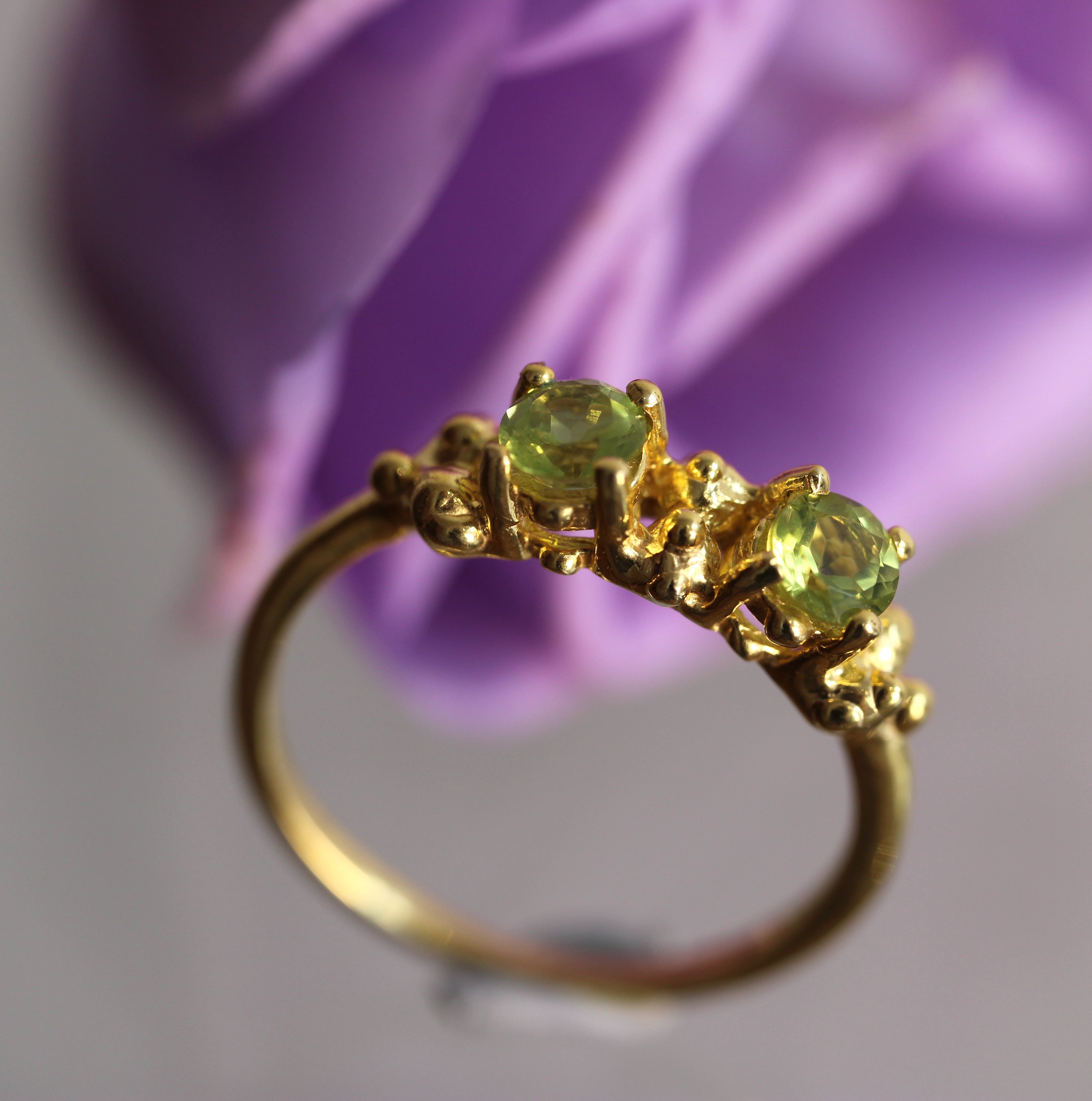 Gold ring with green gemstones against a blurred purple flower background