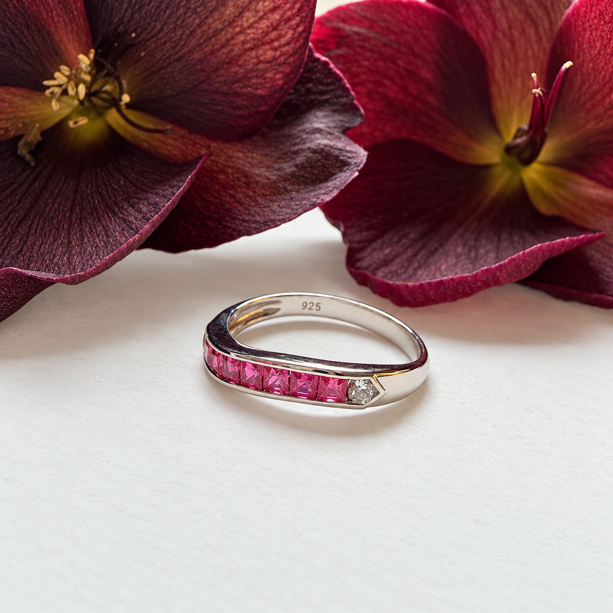 A silver ring with a channel of bright pink gemstones and a small clear stone, photographed on a white surface with dark red flowers in the background.