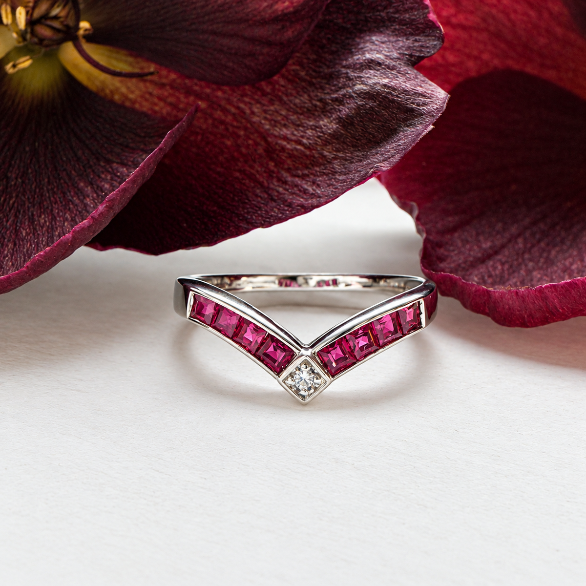 A silver V-shaped ring with bright pink gemstones and a small clear center stone, styled on a white surface with dark burgundy flower petals in the background.