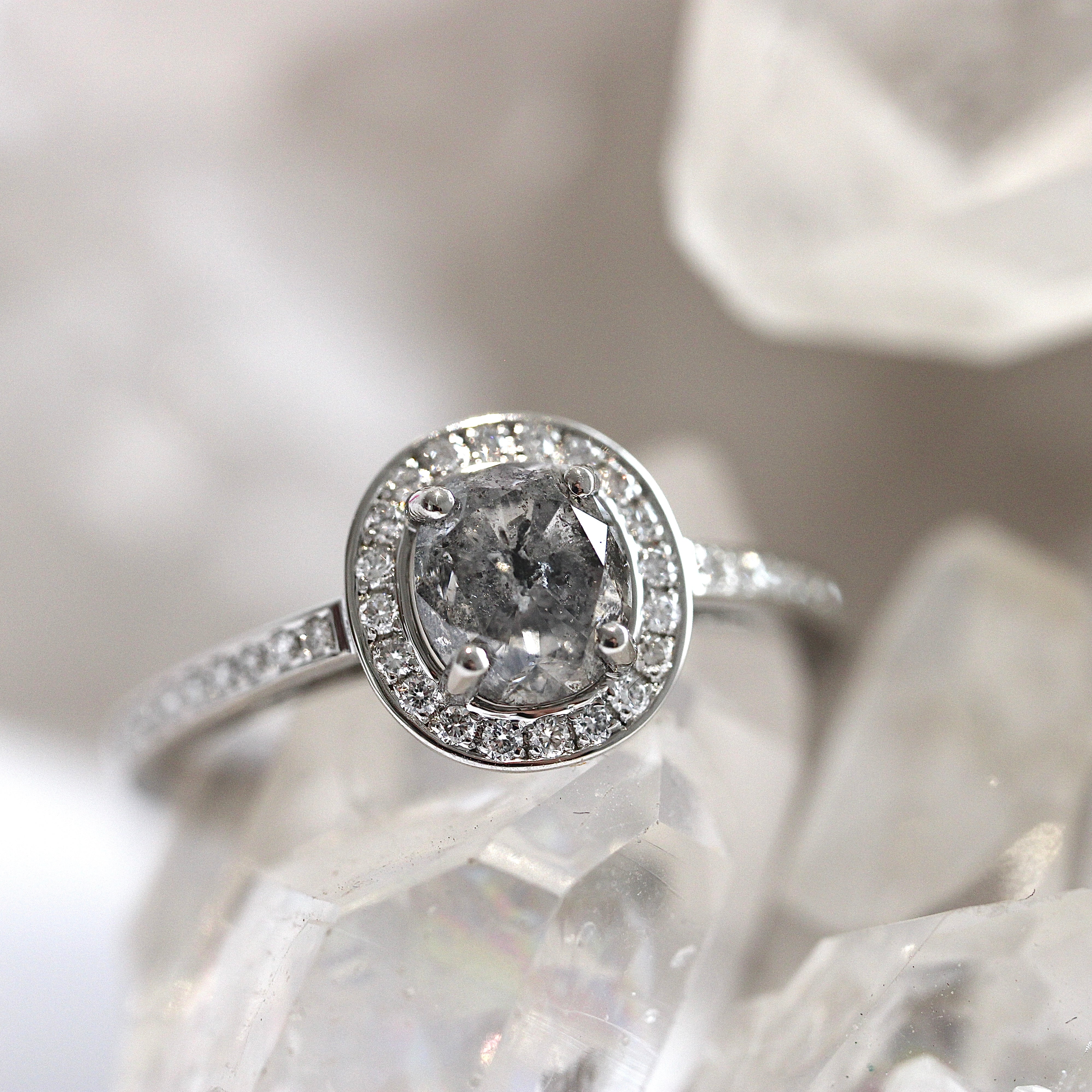 A platinum ring with a central salt and pepper diamond surrounded by smaller white diamond accents on a crystal-clear surface.