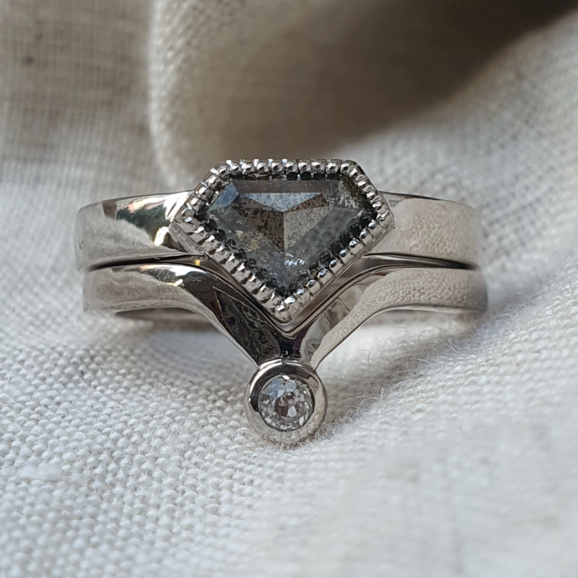 A silver ring with a central hexagonal gray gemstone and a small round clear accent stone, displayed on a textured off-white fabric background.