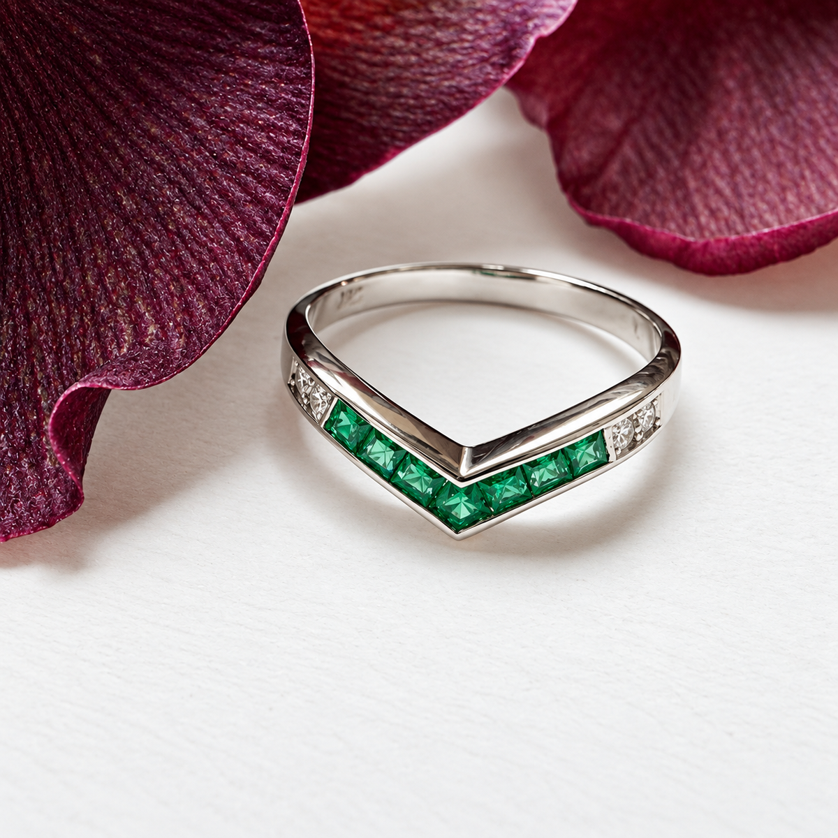 A silver chevron ring featuring a row of green emerald-cut gemstones and small clear accent stones, styled on a white surface with deep burgundy flower petals.