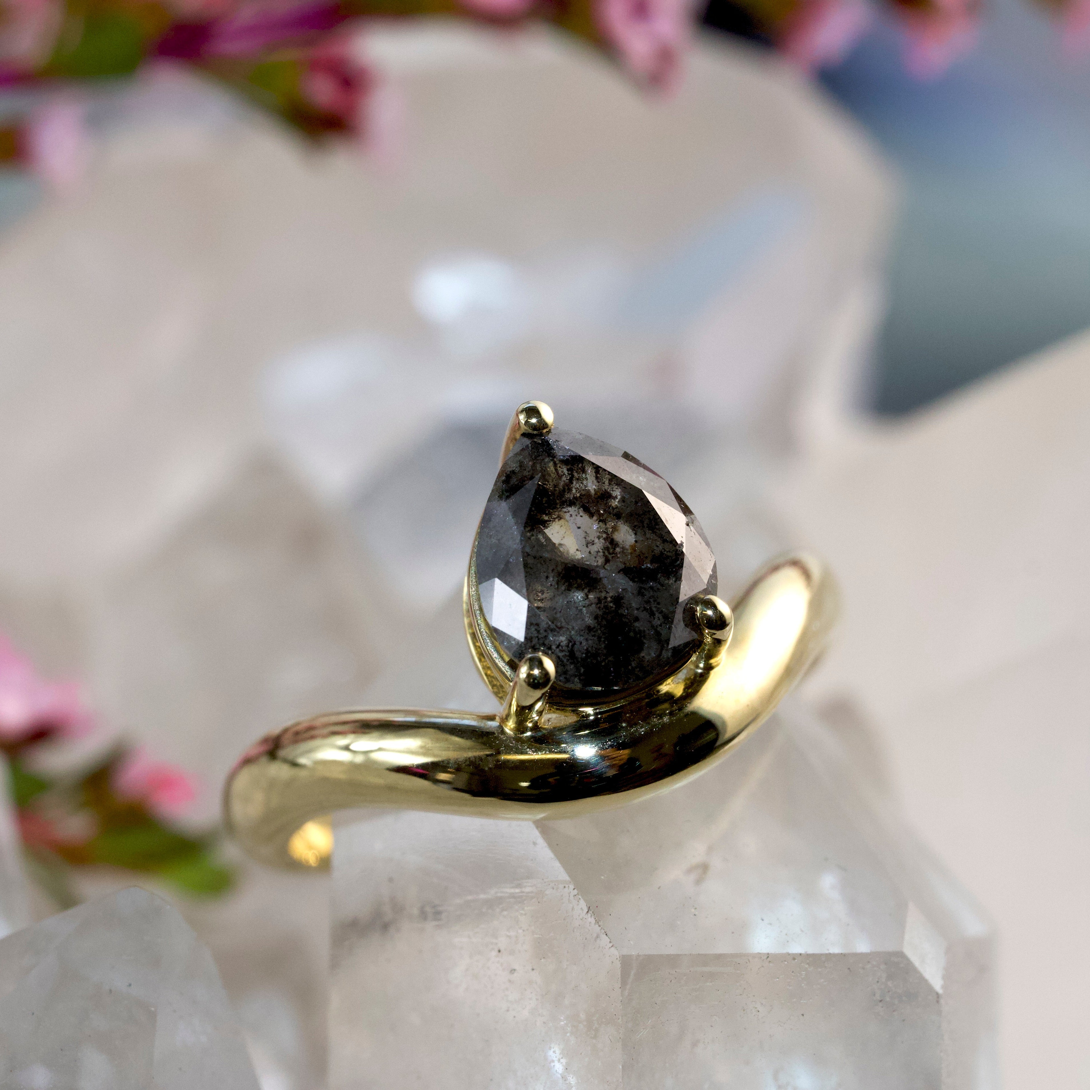 A gold ring with a pear-cut dark gray smoky gemstone, displayed on a clear crystal cluster with soft pink flowers in the blurred background.