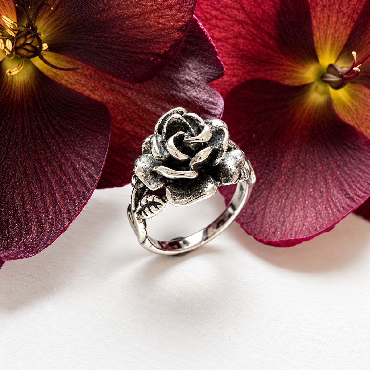 A silver-toned rose ring with detailed petals and leaves, styled on a white surface between deep burgundy flowers.