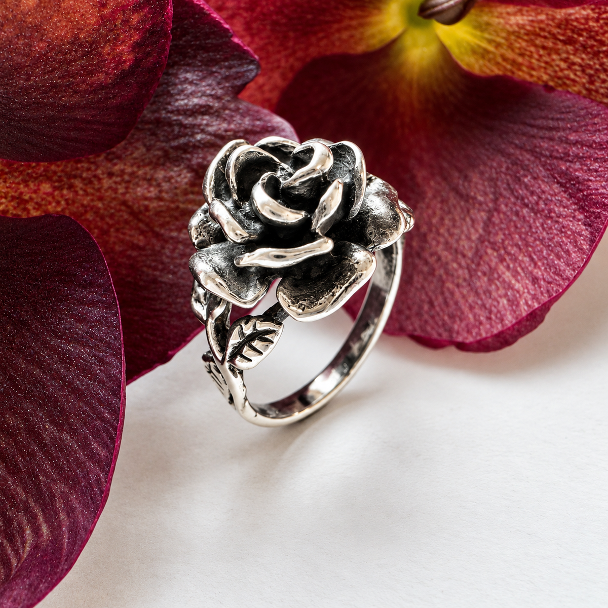 A silver-toned ring shaped like a rose with leaves, displayed on a white surface and framed by large burgundy flower petals.