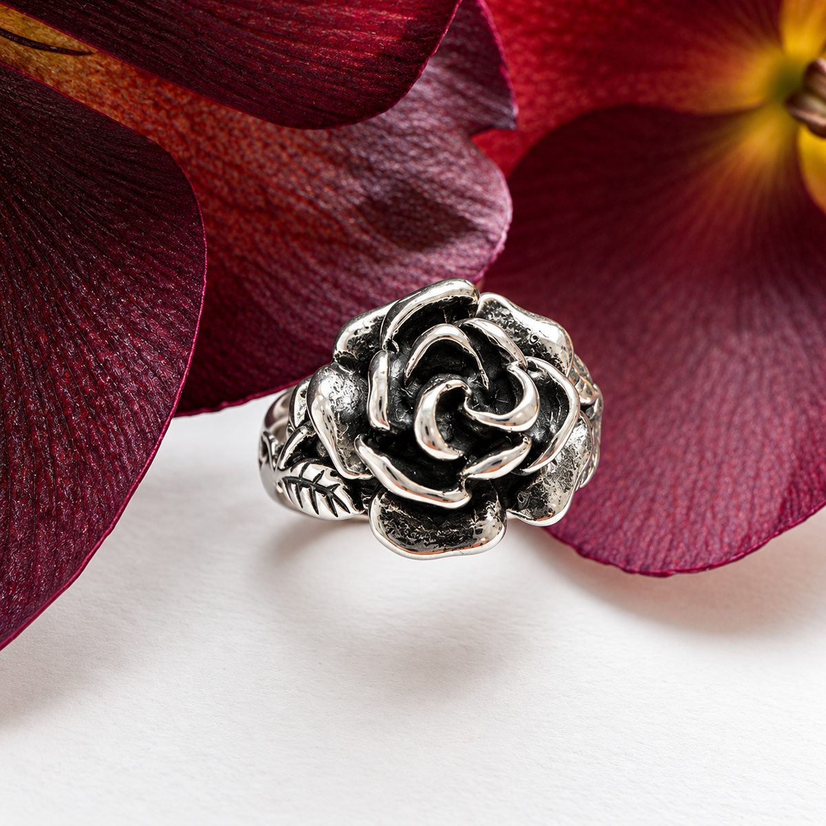 A silver-toned ring sculpted in the shape of a rose, resting on a white surface with large burgundy flower petals in the background.