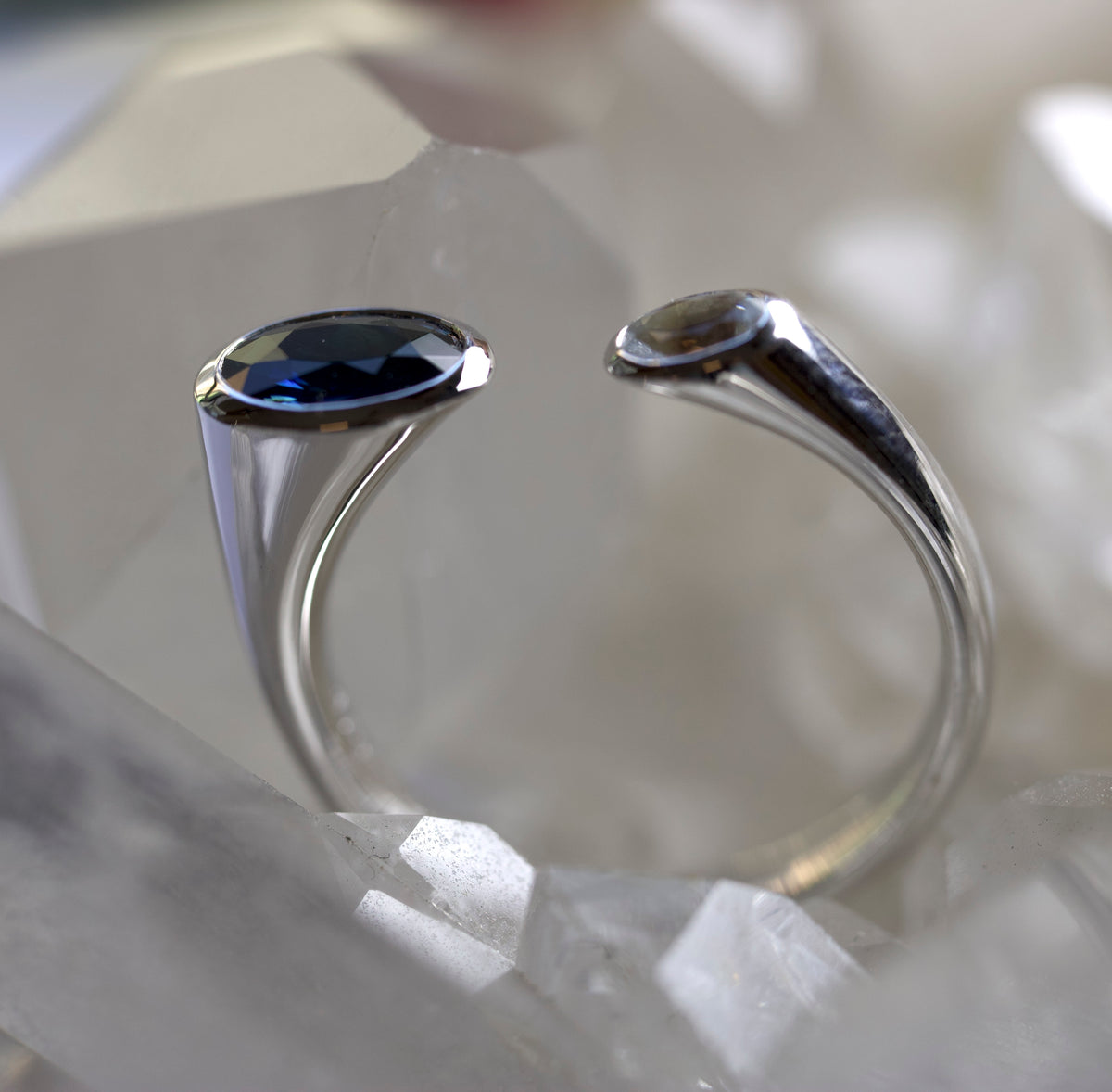 A silver-toned open ring with two oval gemstones, one deep blue and one white sapphire, displayed on white crystal clusters.