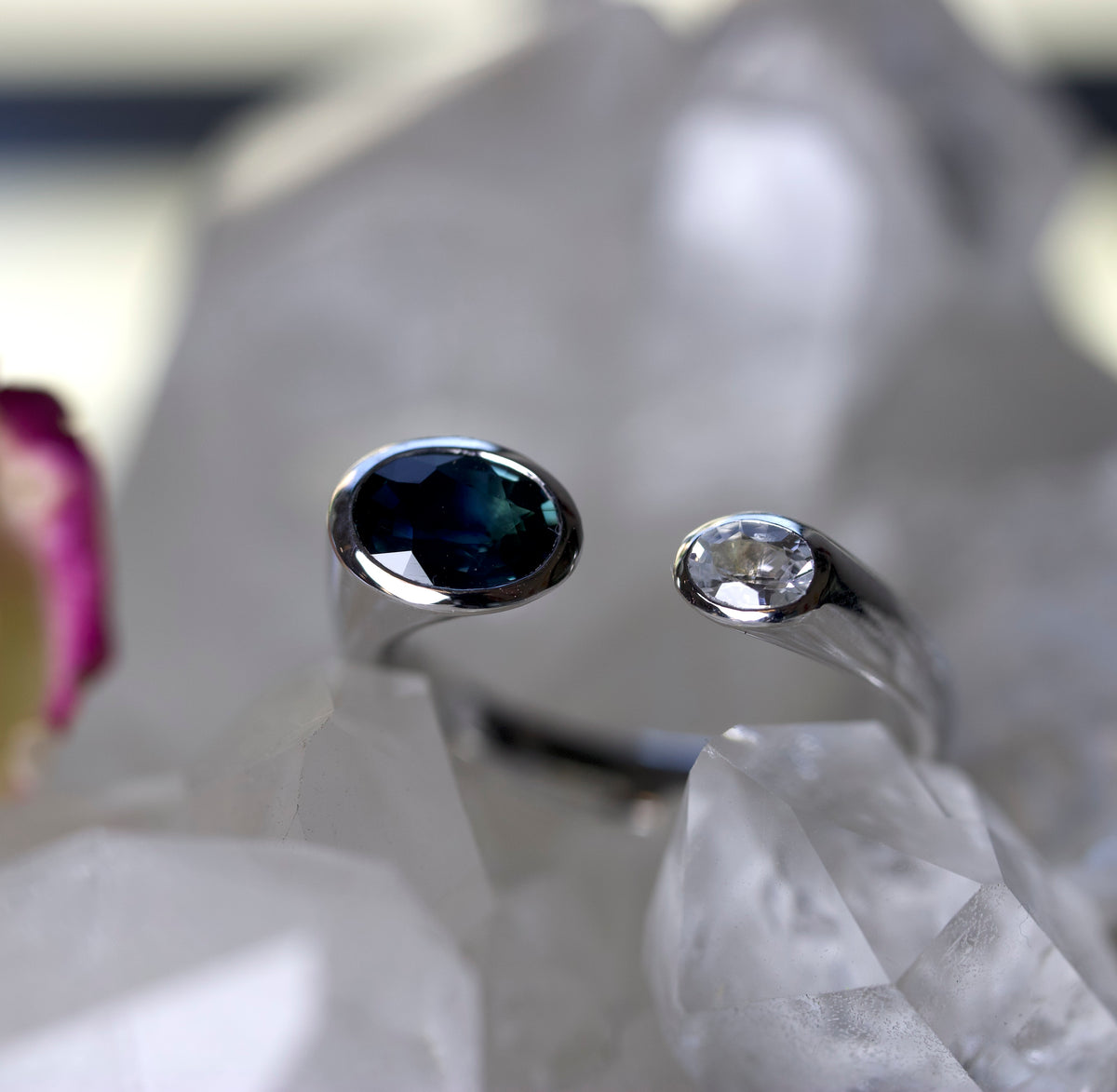 Toi Et Moi silver rings displayed on white crystal points. ring features an oval dark blue sapphire gemstone, and the other features a round clear white sapphire gemstone.