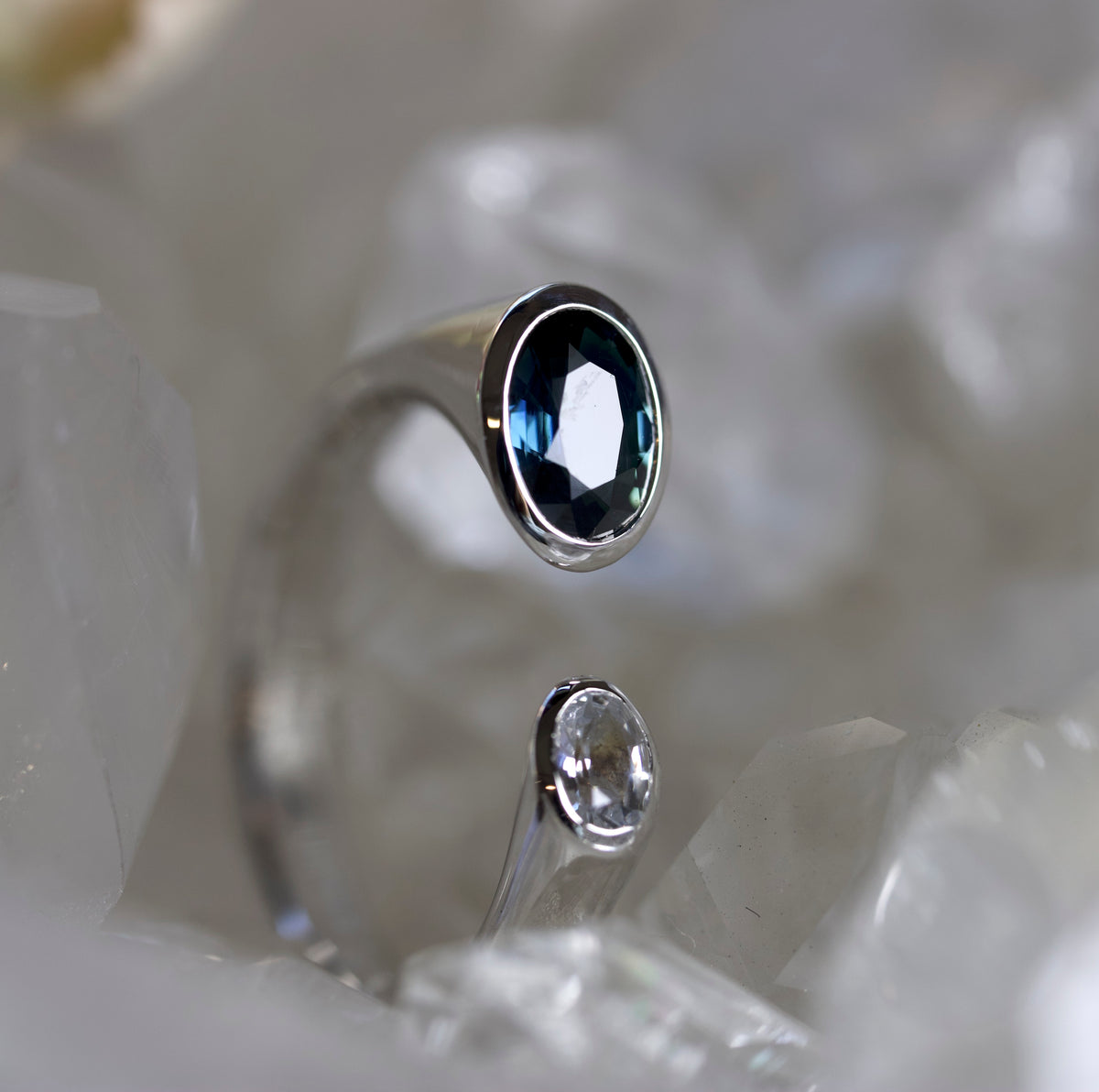 A silver-toned ring with a faceted dark blue oval gemstone, displayed on a bed of white crystal formations. A smaller, clear gemstone version of the same ring is reflected below it.