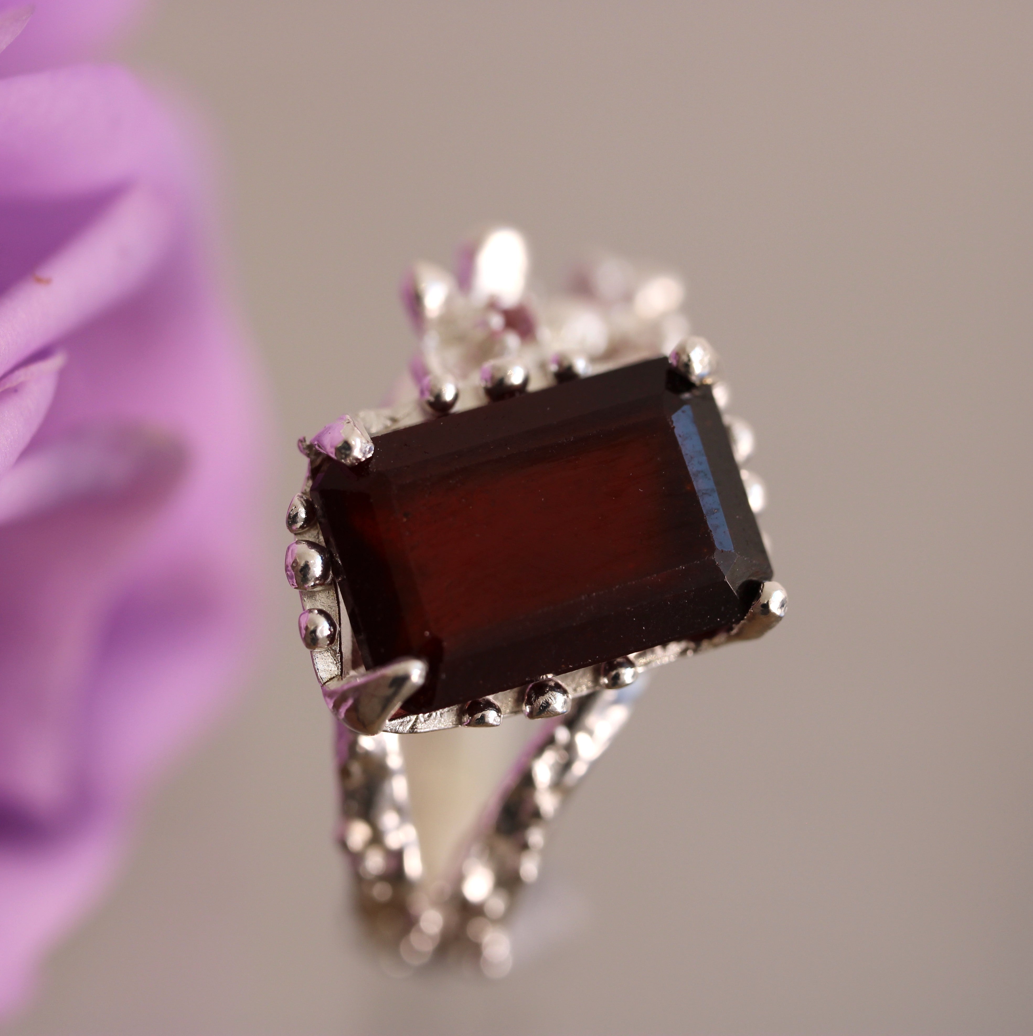 Silver ring with a rectangular red gemstone against a blurred purple flower background