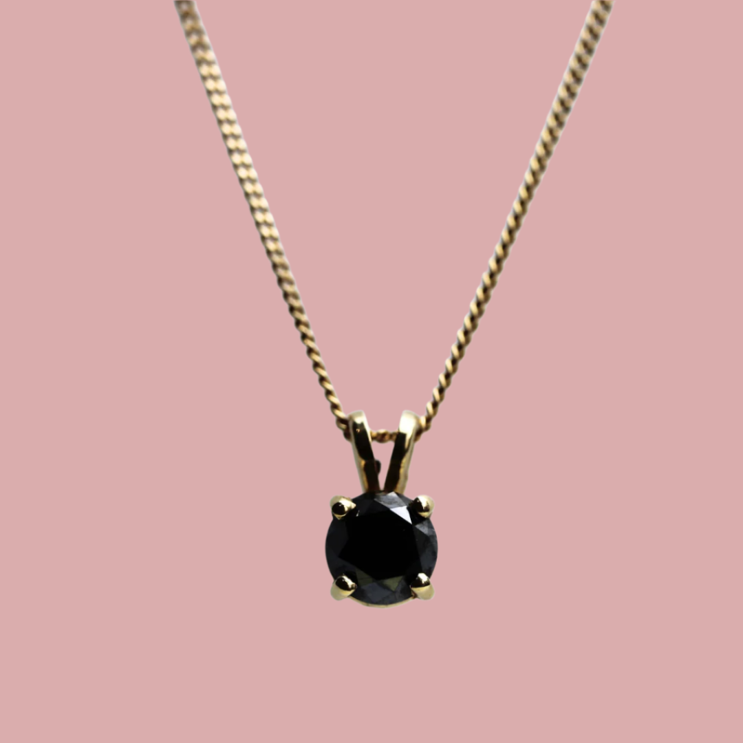 A black diamond pendant set in a gold setting, displayed on a pink background.