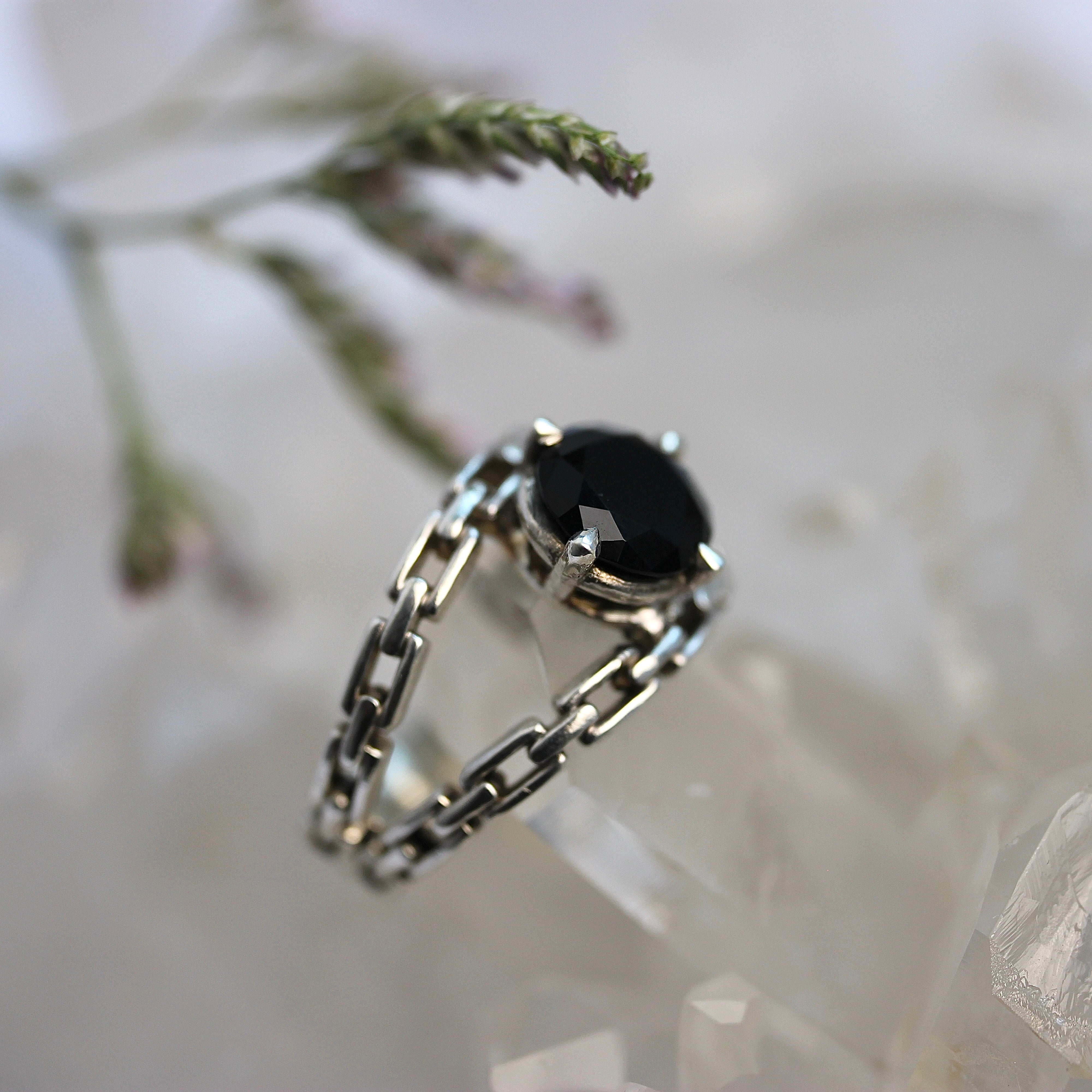 Silver chain ring with a black gemstone on a crystal surface with blurred plant background