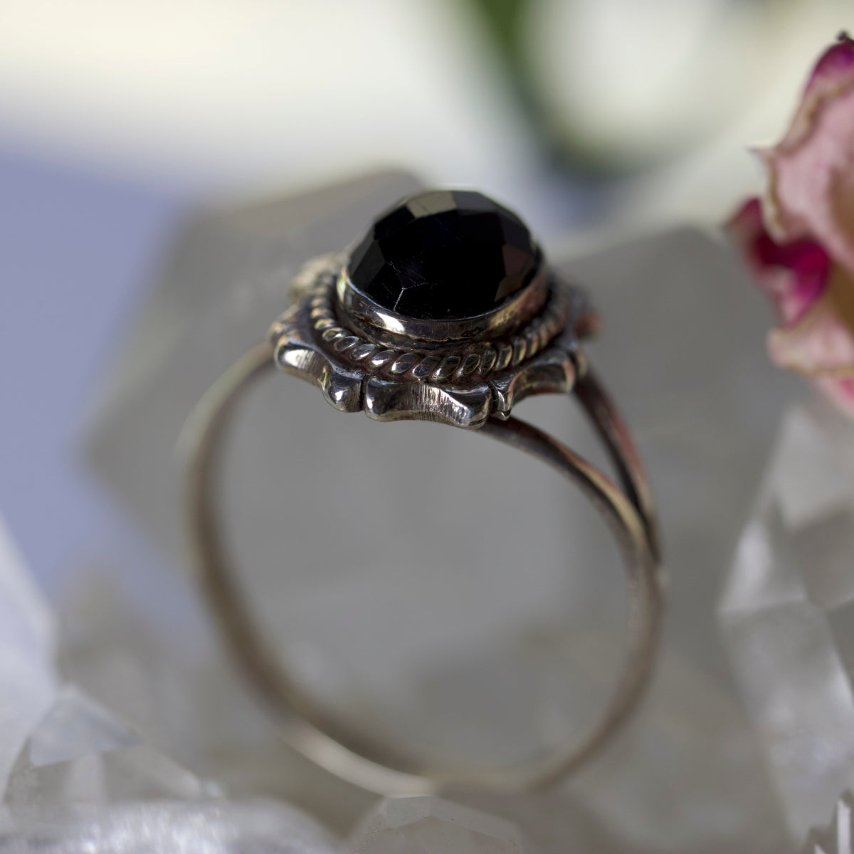 A silver-toned ring with a faceted black gemstone, displayed on a clear crystal stand with a soft-focus background.