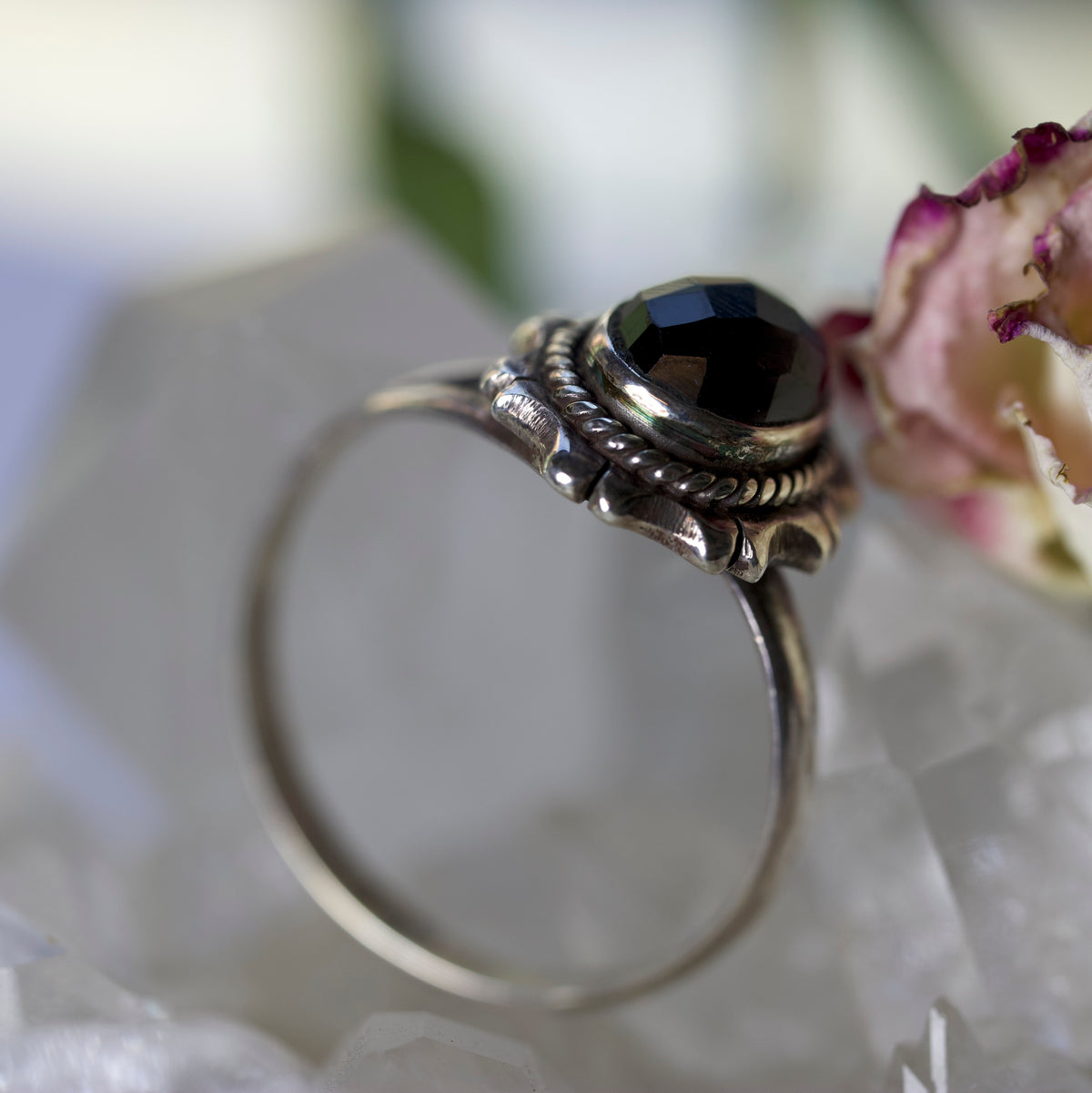 A silver-toned ring with a faceted black gemstone and ornate textured detailing, resting on white crystal formations with a blurred dried flower in the background.