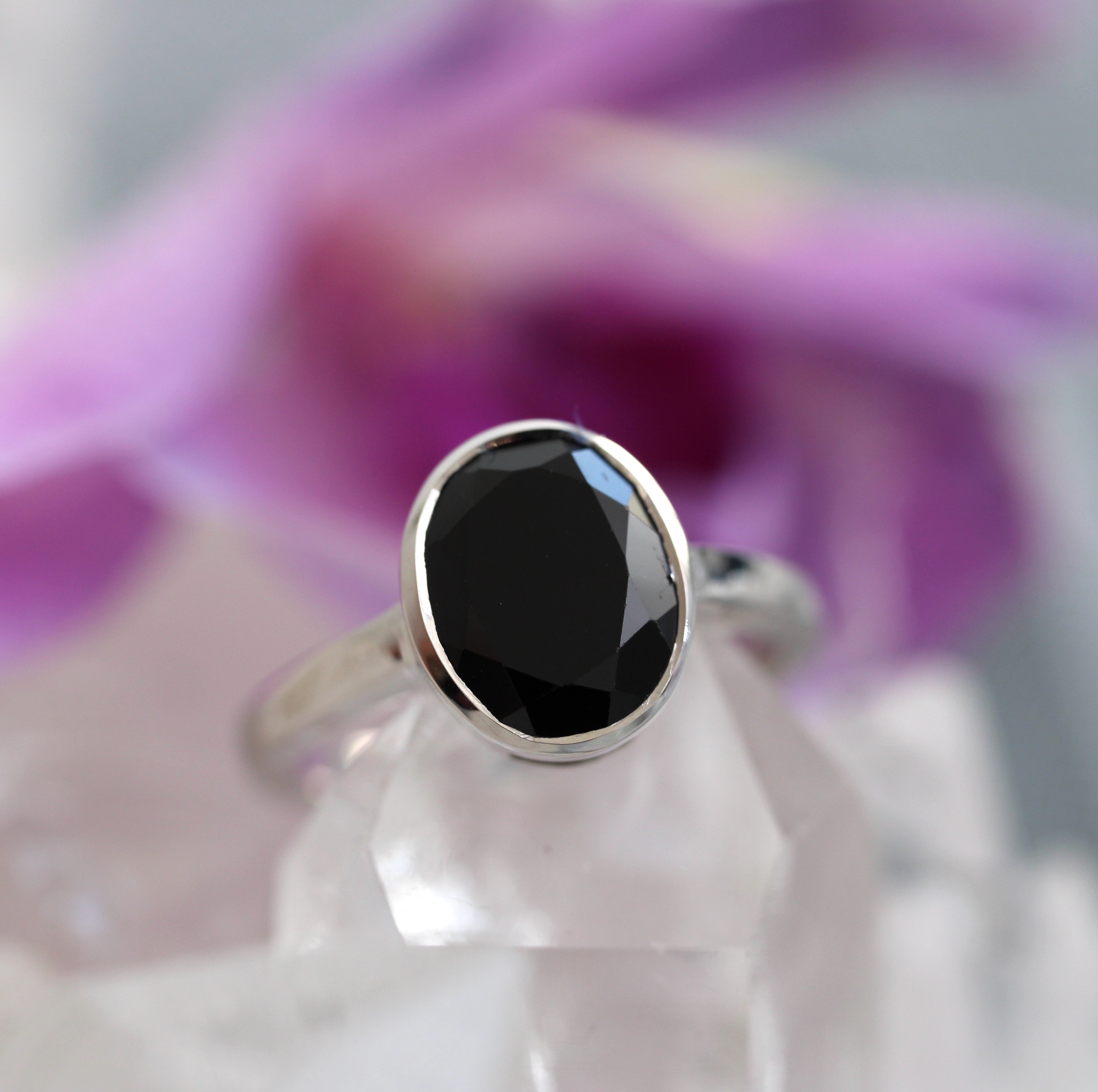 Ring with a black tourmaline on a white stand against a blurred purple flower background