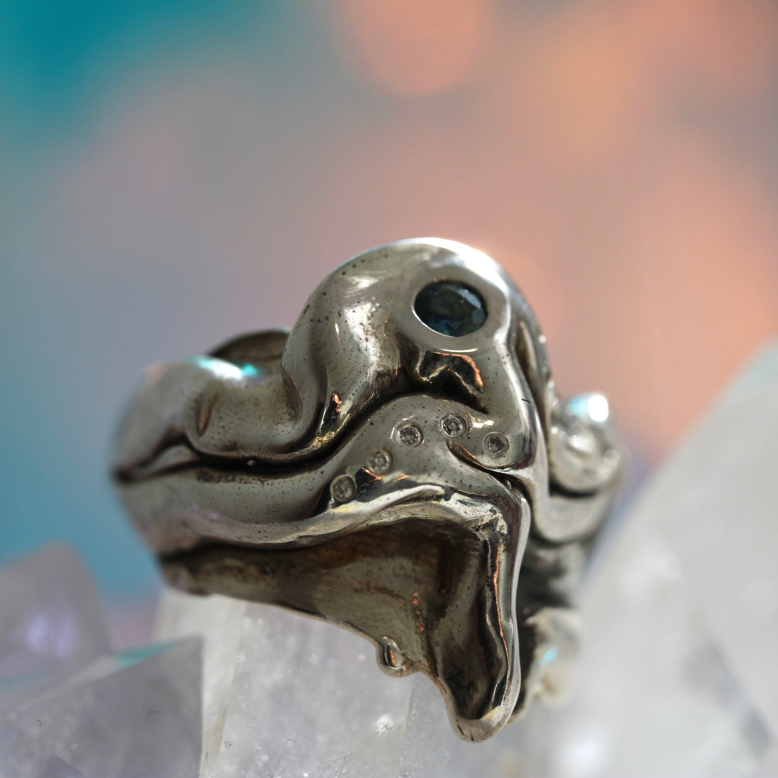 A close-up image of a handcrafted sterling silver ring with textured details and adorned with small salt + pepper diamonds and a blue partii sapphire.