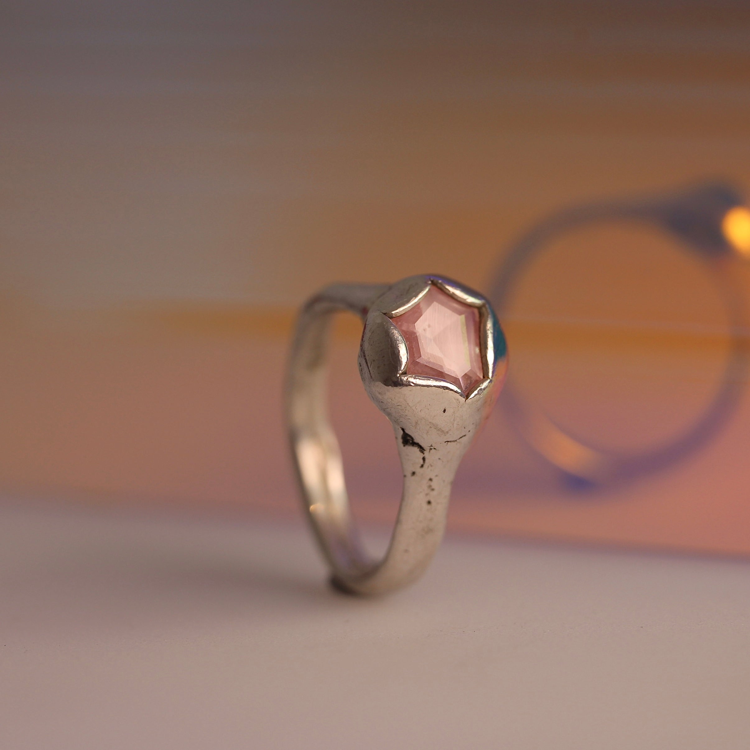 A sterling silver ring with a geometric cut pink sapphire set in a textured setting, displayed against an orange and pink background.