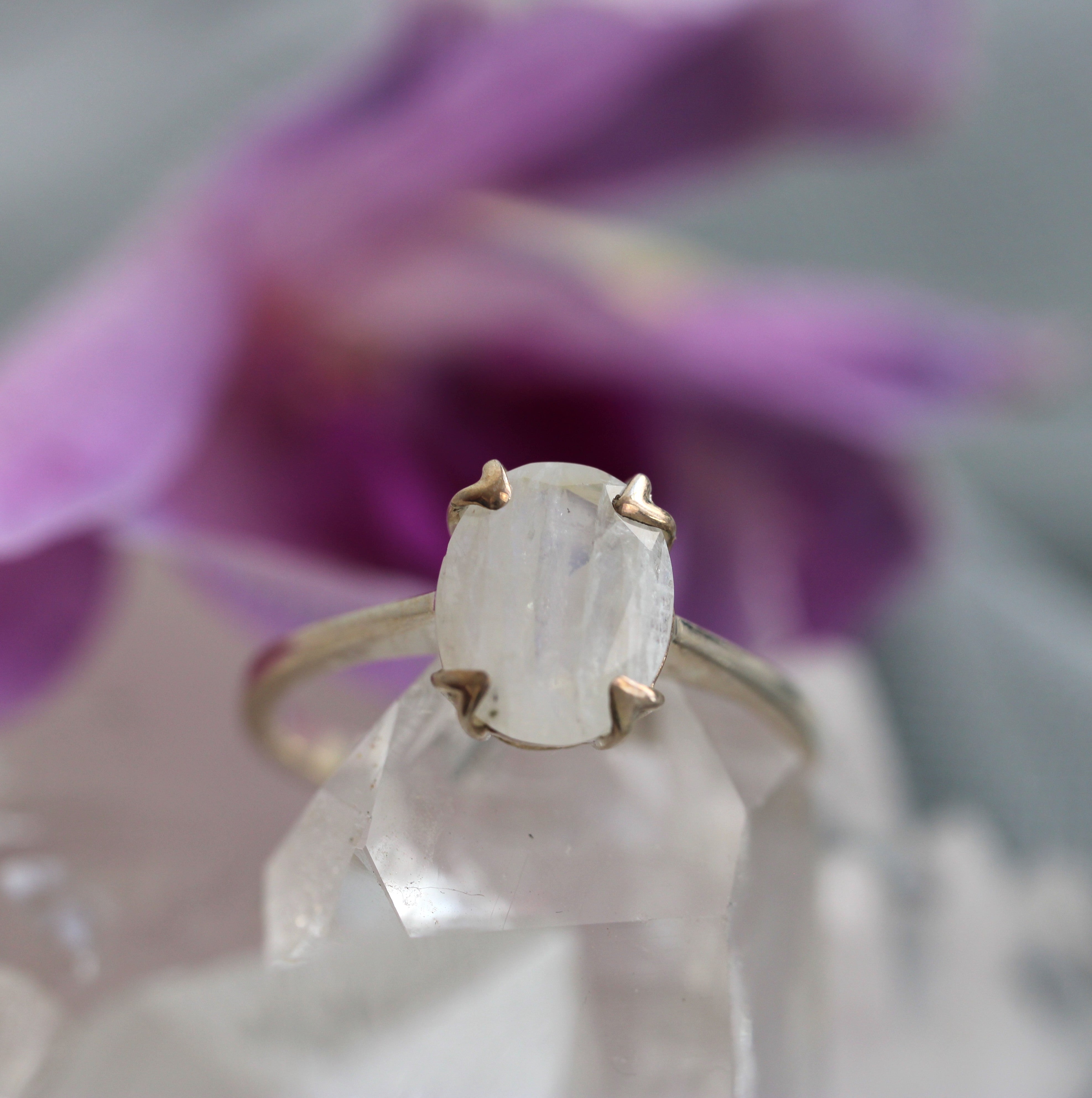 Gold ring with a central gemstone on a clear crystal with a blurred purple flower in the background