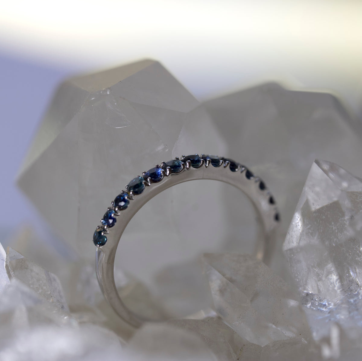 A silver band ring set with small dark blue round gemstones, resting on clear crystal clusters against a bright white background.
