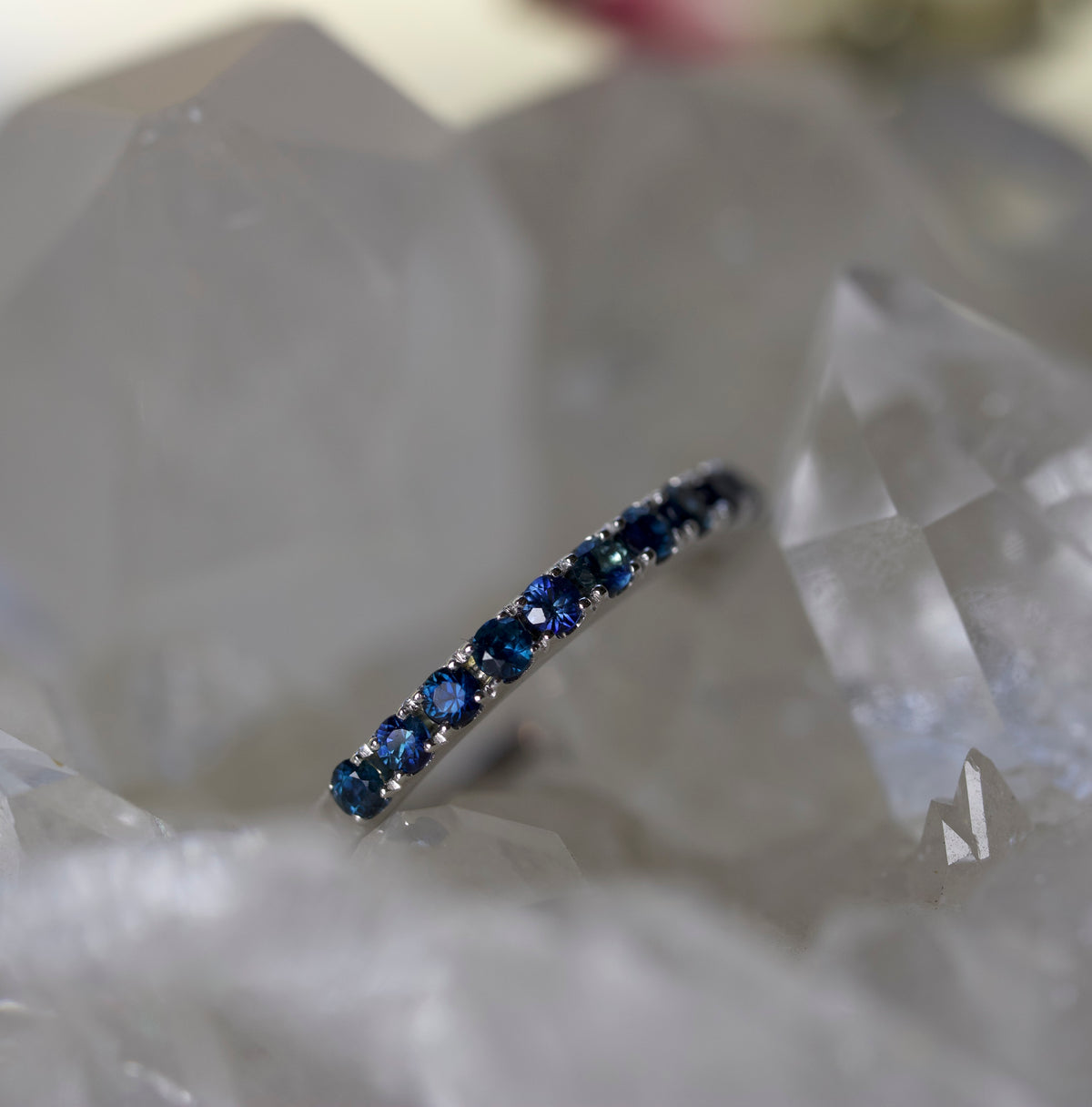 A slim silver band ring set with a row of deep blue round gemstones, resting on a bed of white crystal points.