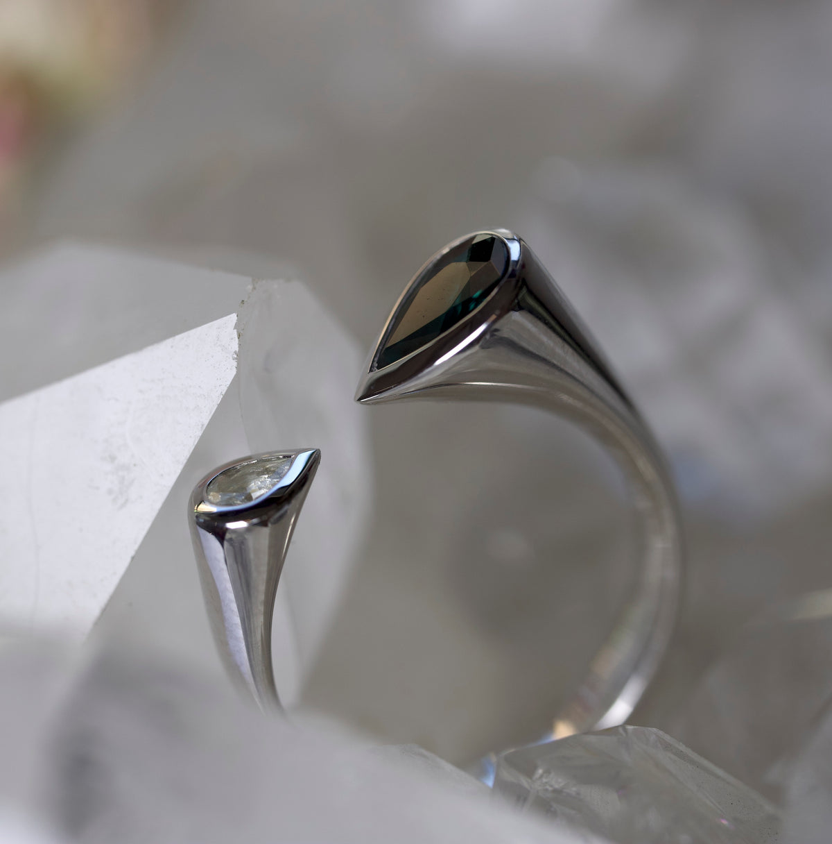 A silver open ring with two teardrop-shaped settings, one holding a clear gemstone and the other holding a dark iridescent stone, displayed among translucent white crystal points.
