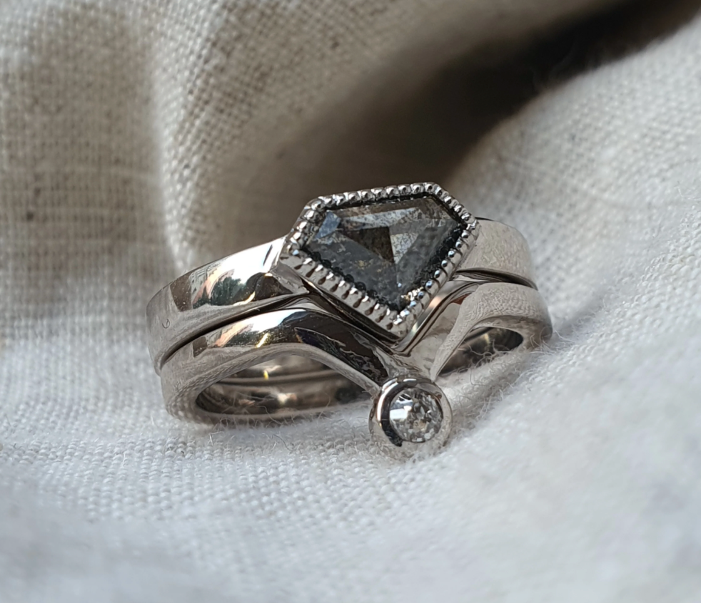 A silver-toned ring set featuring a central triangular dark gray gemstone in a beaded halo setting, with a smaller clear round gemstone on the side, displayed on a textured off-white fabric background.