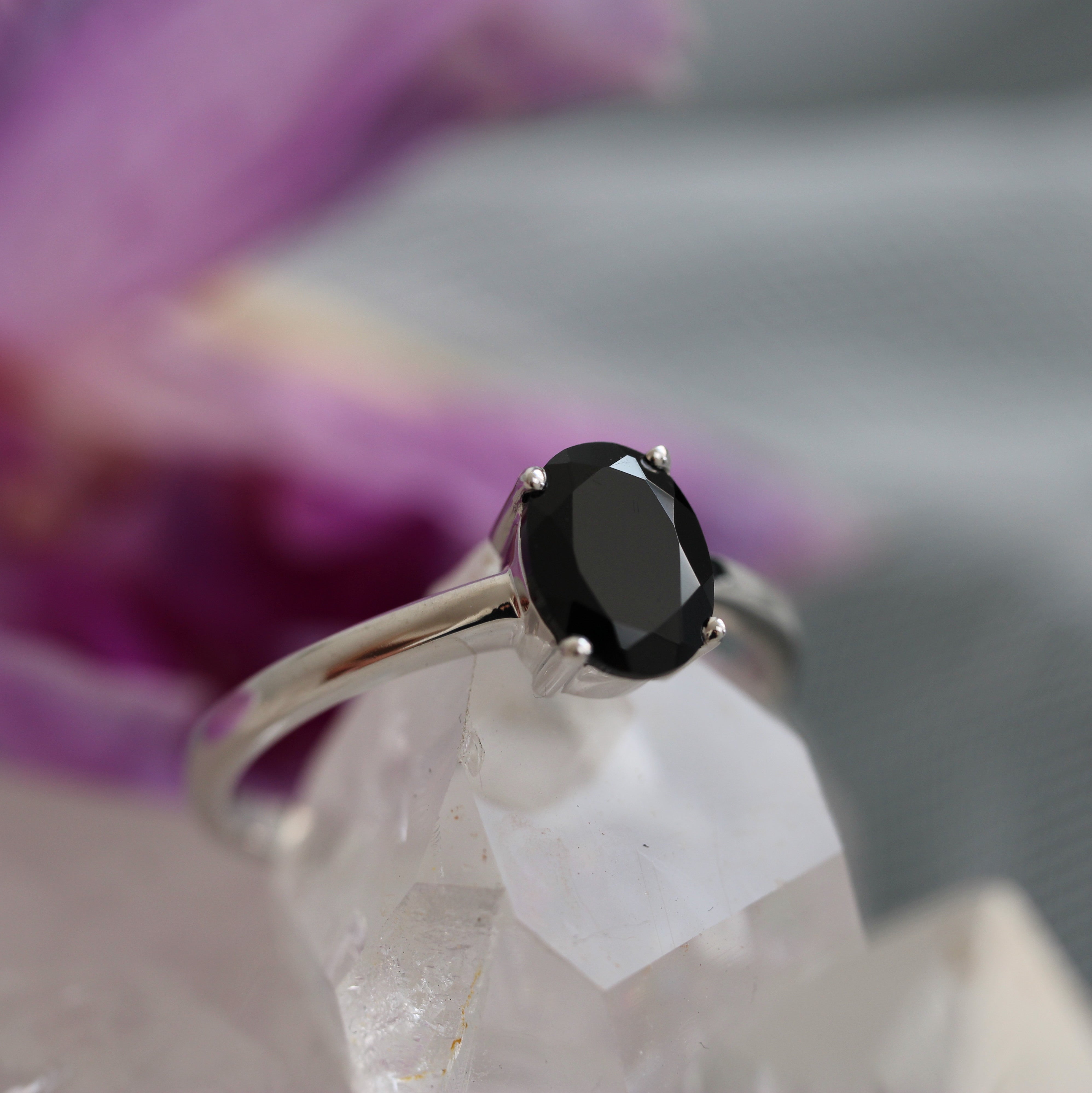 Silver ring with a black gemstone on a clear crystal surface with blurred purple flowers in the background