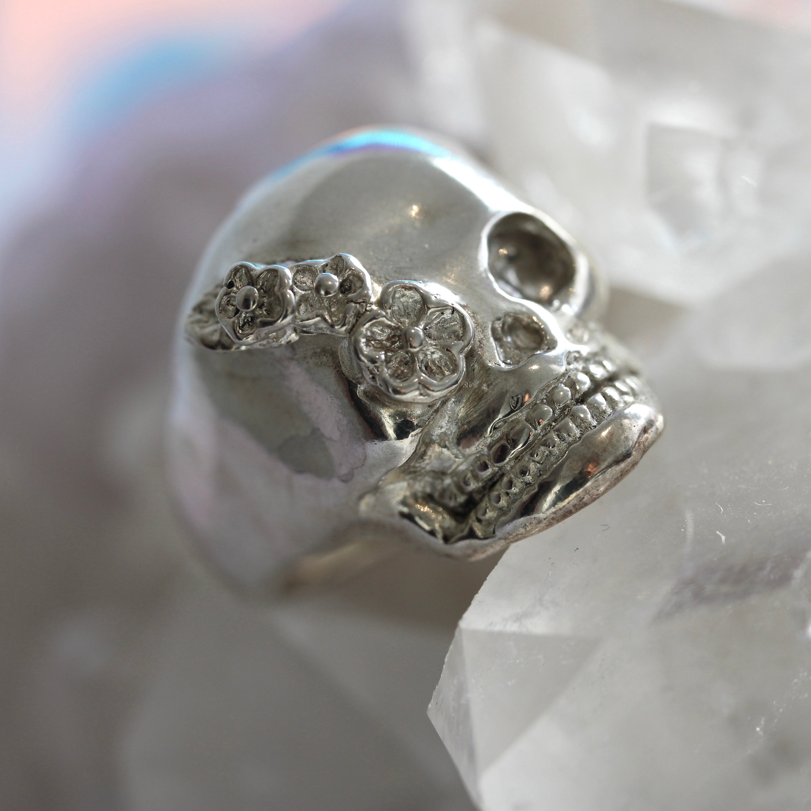 A solid sterling silver skull ring with floral accents on the face, displayed against a crystal background.
