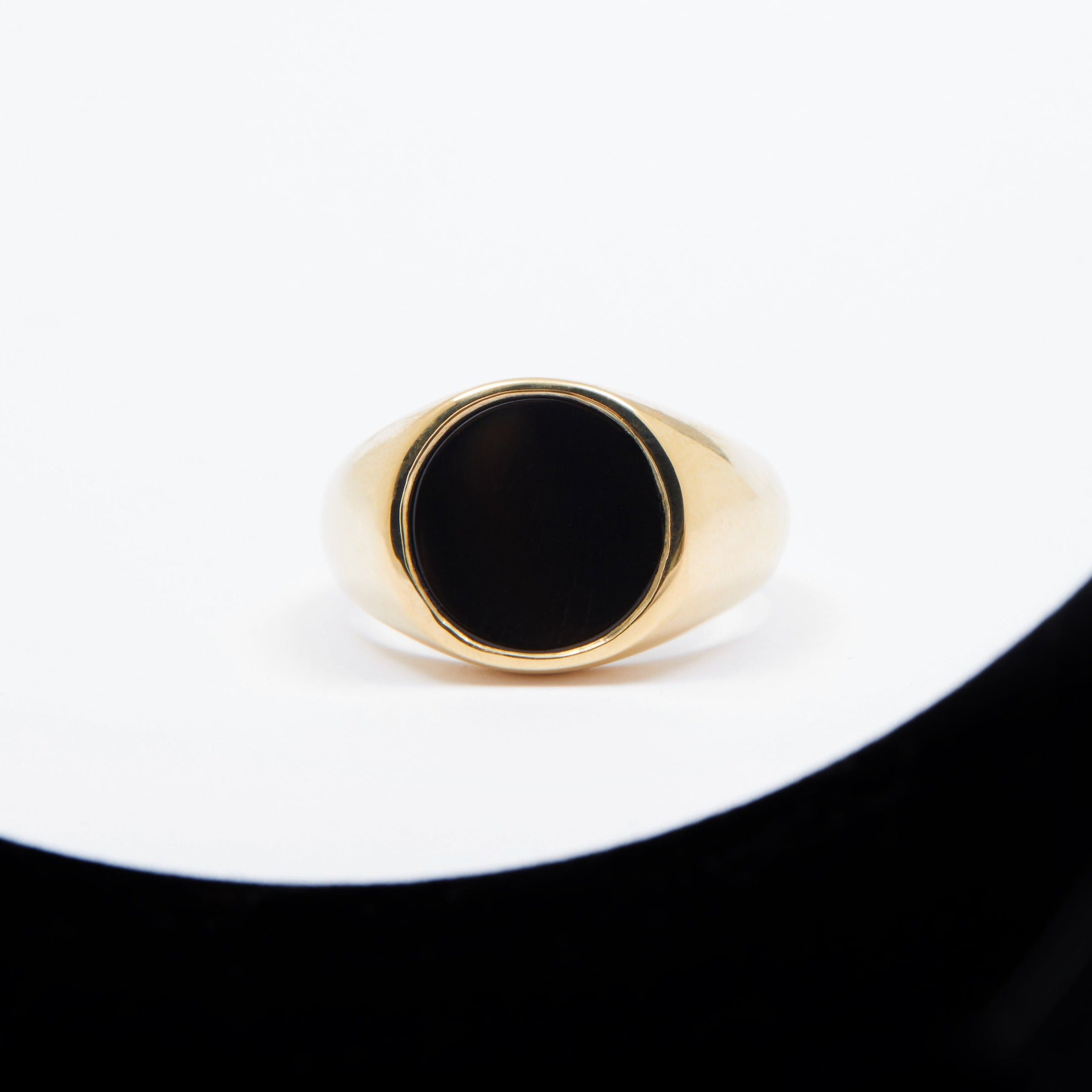A signet ring with a round black onyx inlay set in a gold band, displayed on a white and black surface.