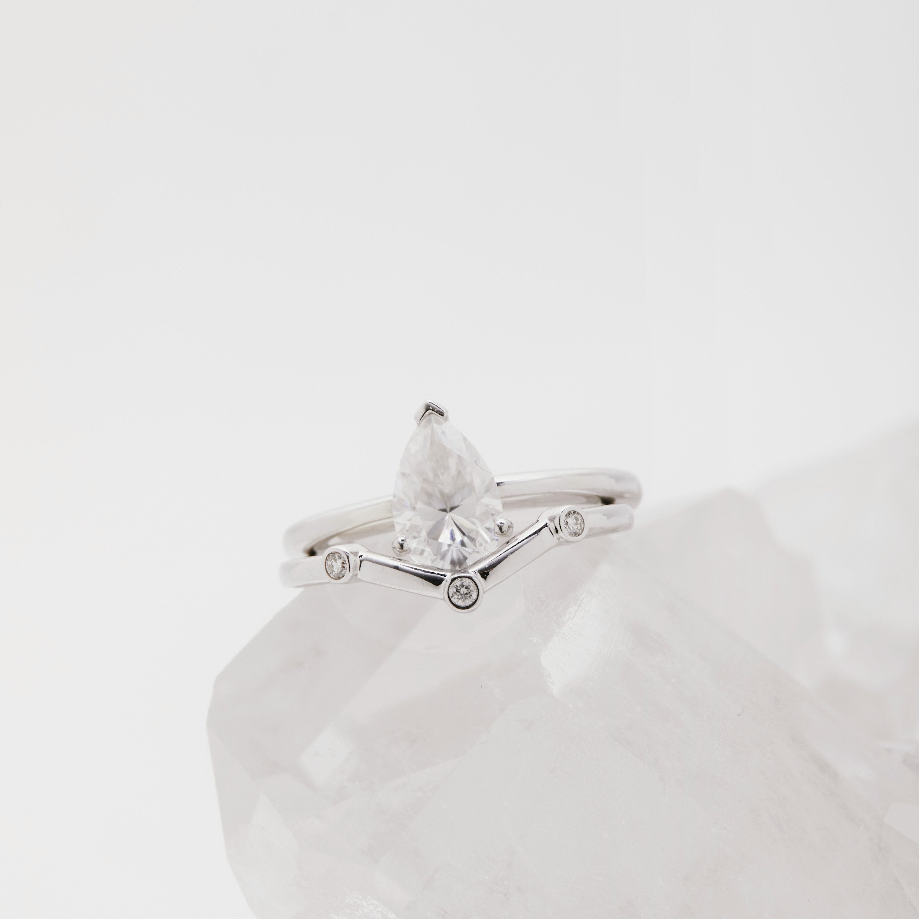 A silver-colored chevron shaped wedding band with three small flush-set diamonds, displayed on a clear glass surface.