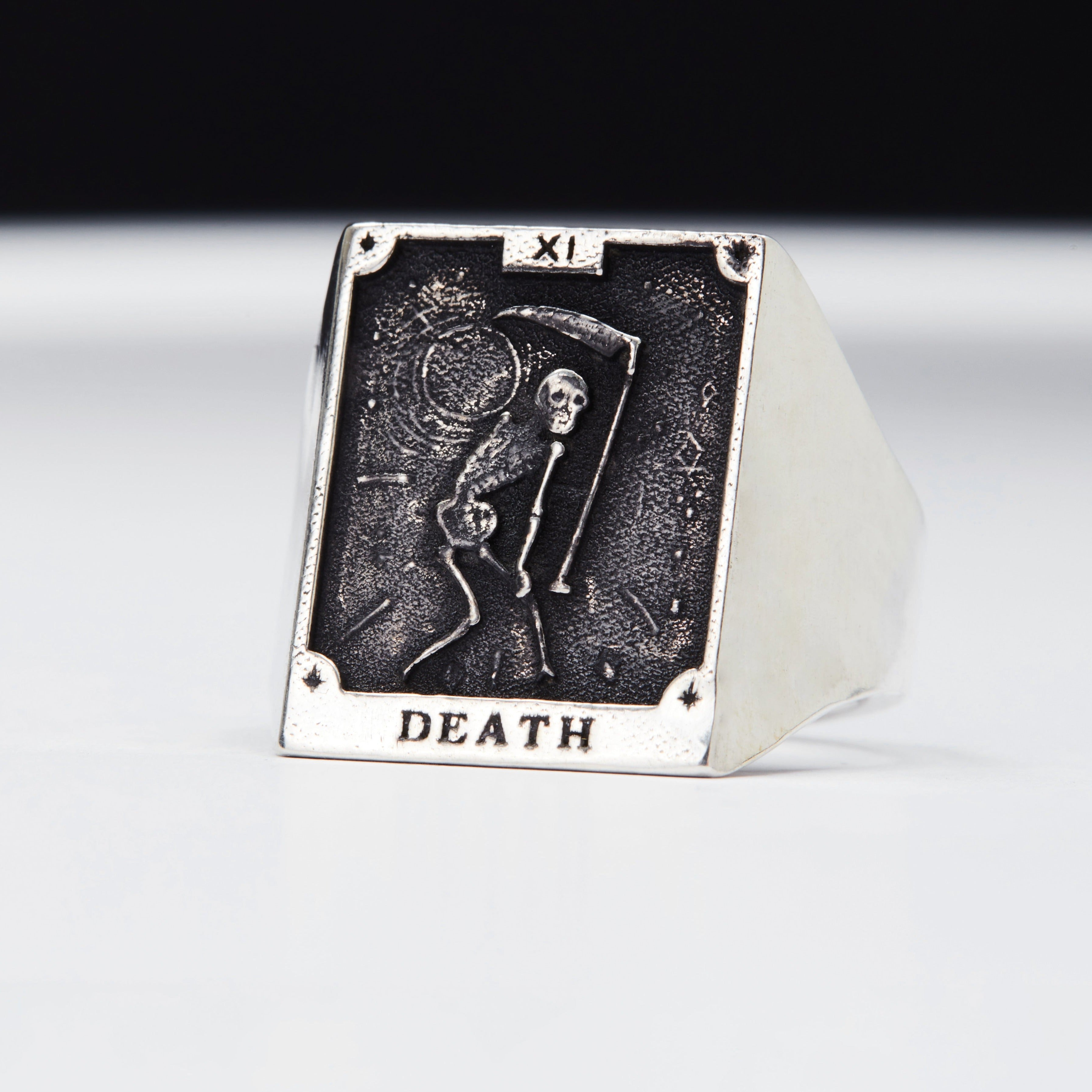 A signet ring featuring a tarot card motif with the word 'DEATH' written on the card, set against a black background. The ring is made of a precious metal, possibly silver, and has a skull and crossbones emblem.