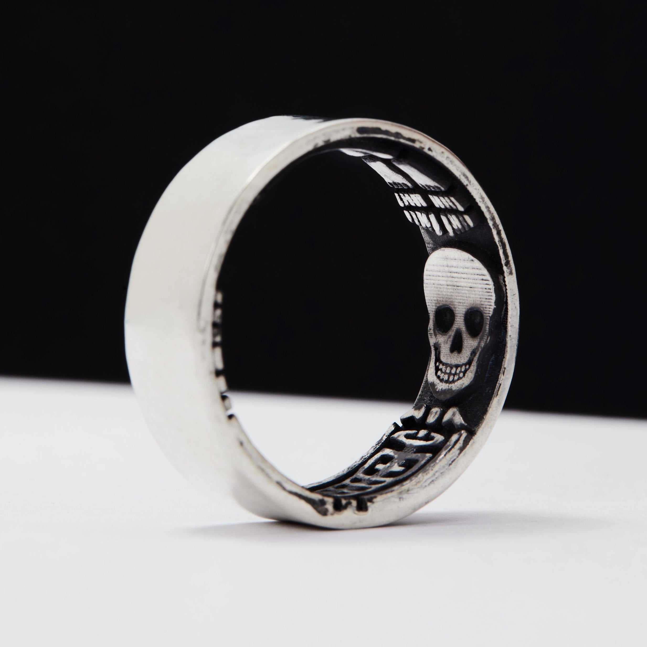 A flat wedding band with a memento mori design, featuring a blackened skull in the center, oxidised for a black and silver contrast, with a textured background.