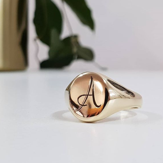 A gold signet ring with an engraved letter 'A' on a white surface with a plant in the background.